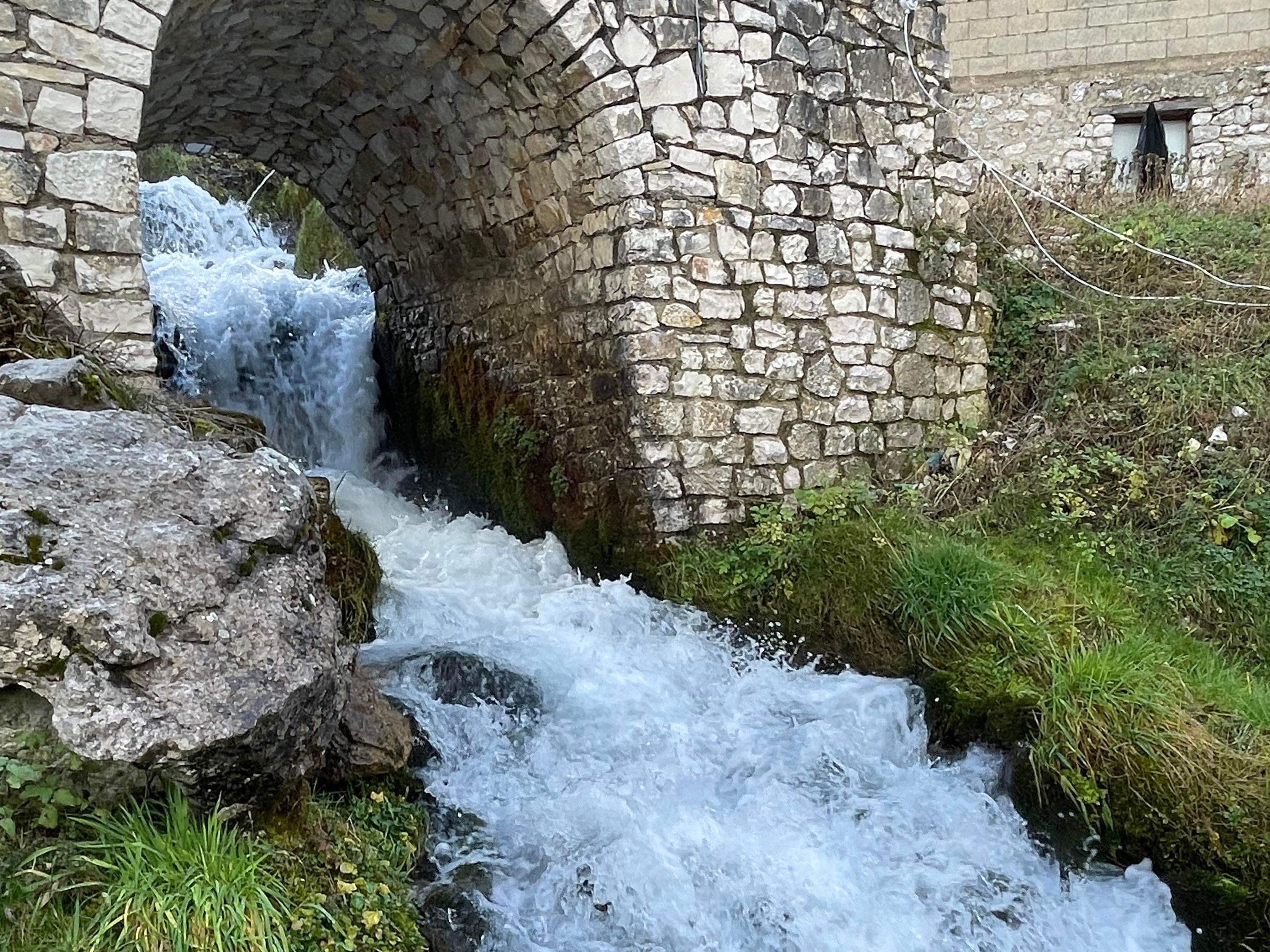 A water stream flows under an arched stone bridge, surrounded by lush grass and rocks.