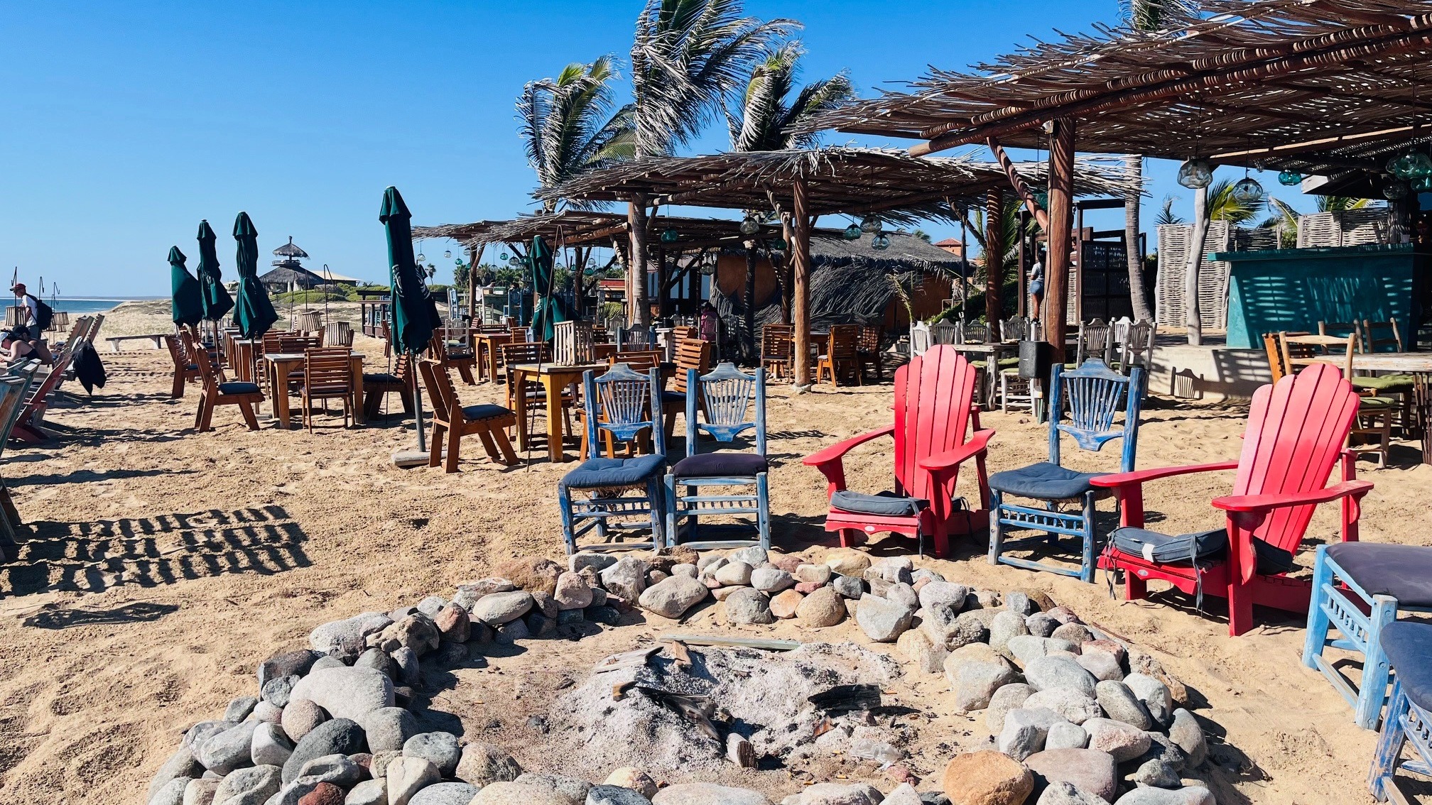 A beachside seating area with chairs, tables, a fire pit under palm trees, and a clear blue sky.