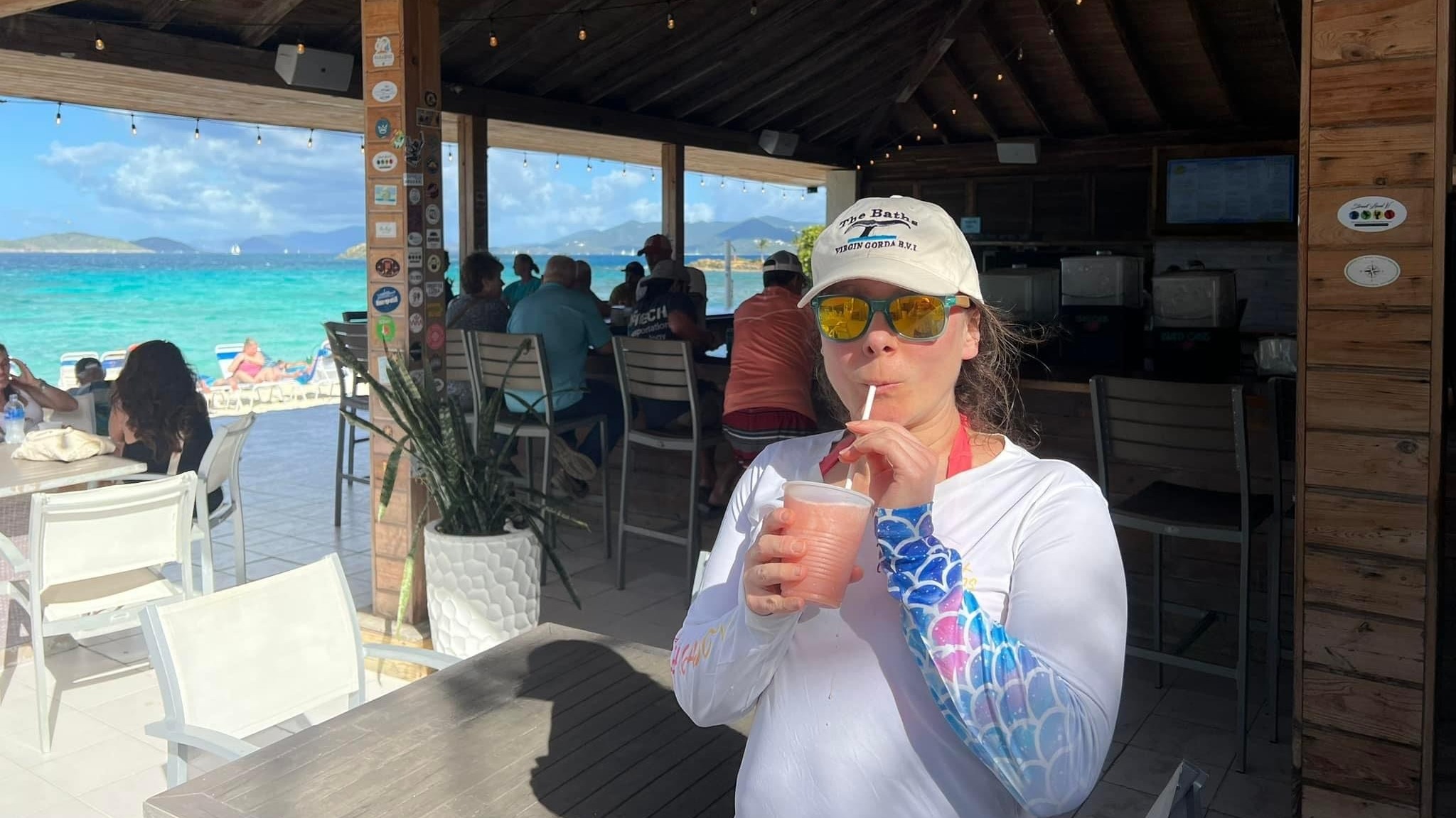 A woman in sunglasses and a casual beach outfit enjoys a drink at an open-air bar with a stunning turquoise ocean view in the background.