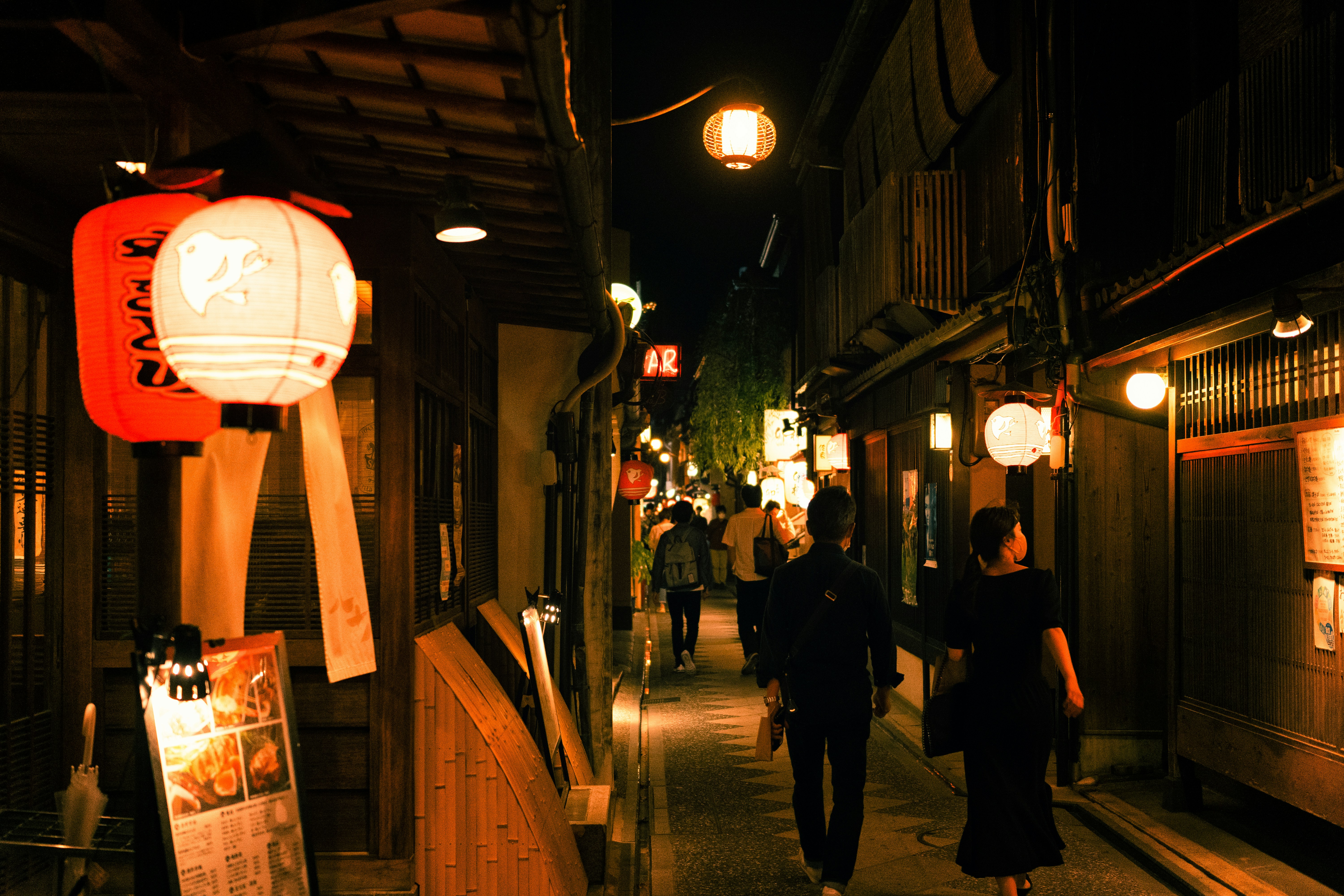 The image shows people walking at night down a narrow alley illuminated by Japanese lanterns and street lamps.