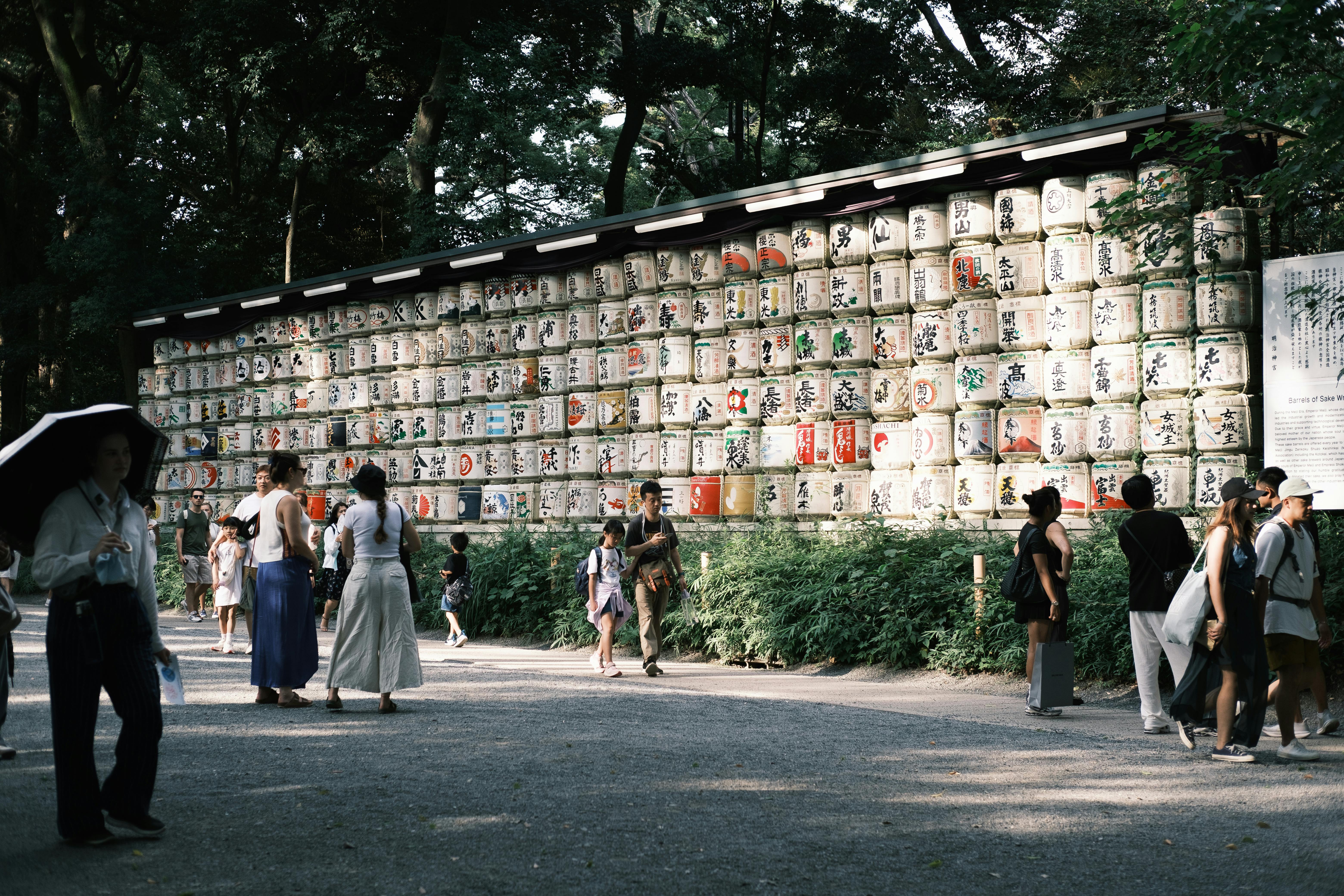The images shows people walking leisurely along a path in front of a large structure holding many rows of white barrels with Japanese artwork.