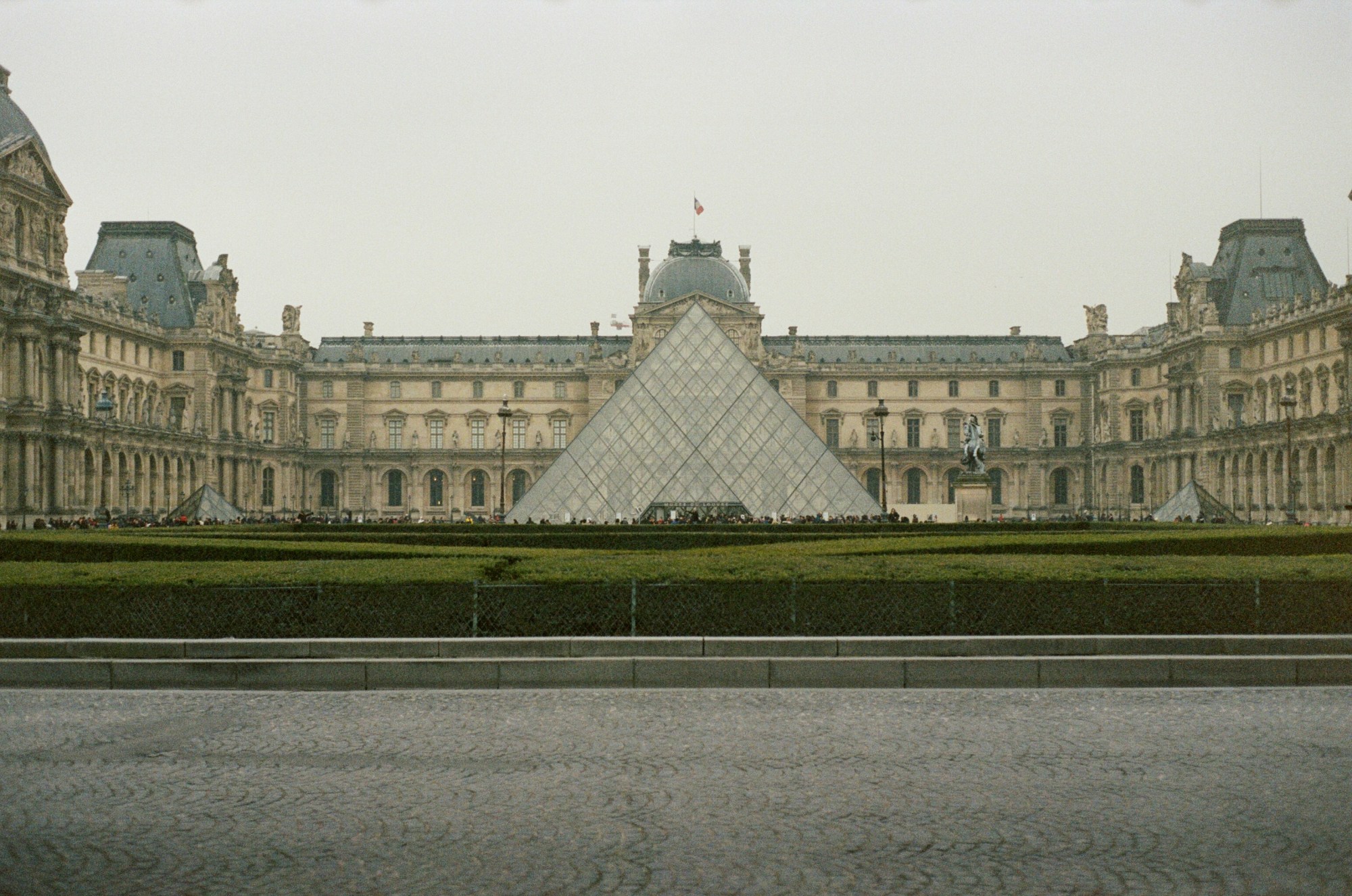 A large building with historical architecture and a pyramid structure in the front and center on an overcast day.