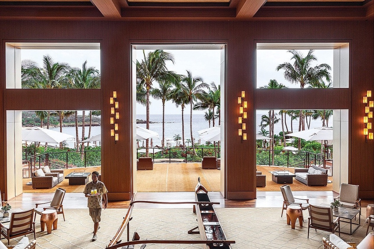 looking out toward sea and palm trees from inside a hotel open-air lobby with tall ceilings.