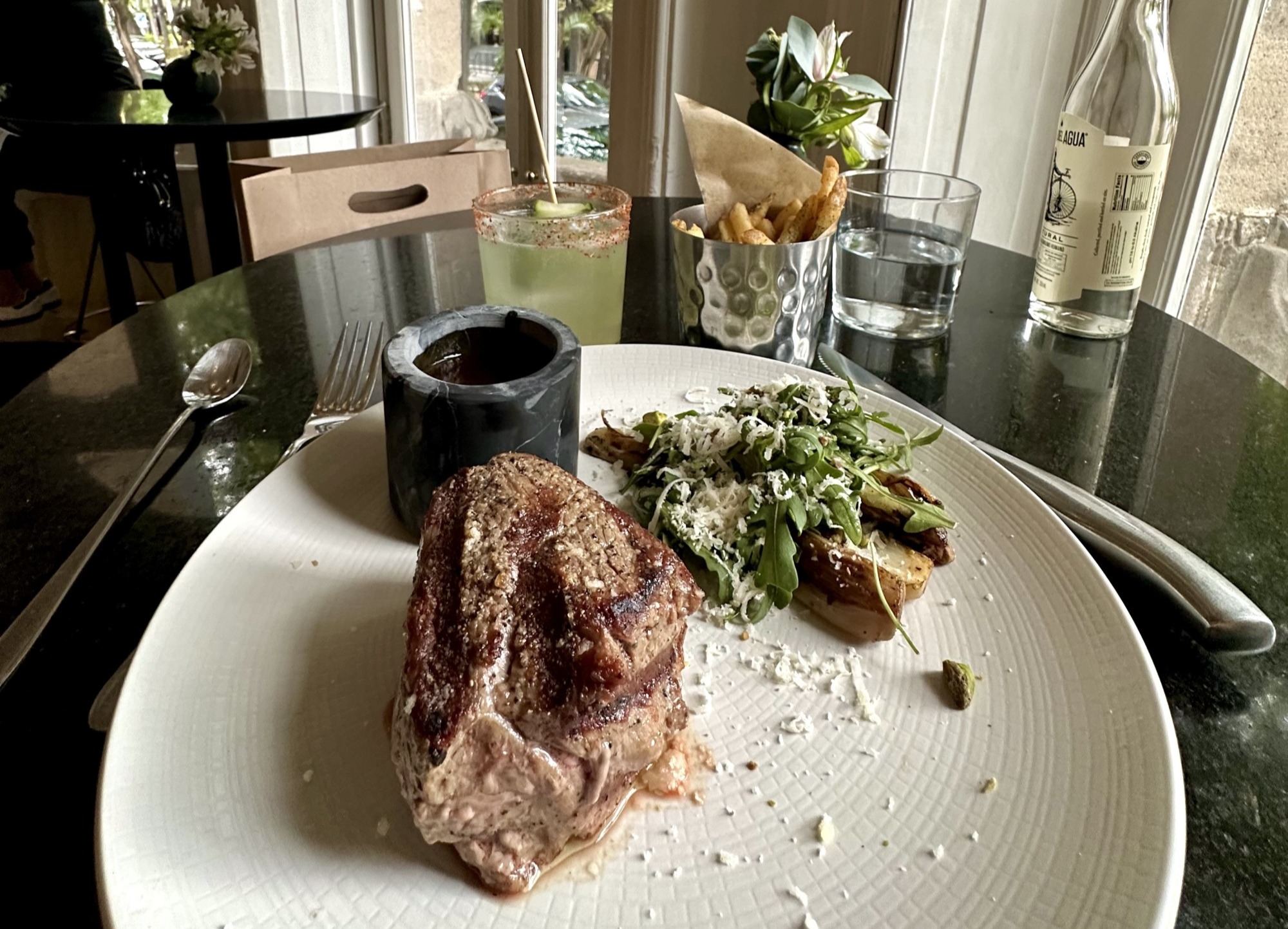 A food view of a large piece of steak with greens on a white plate and a glass and bottle of water on a black table.