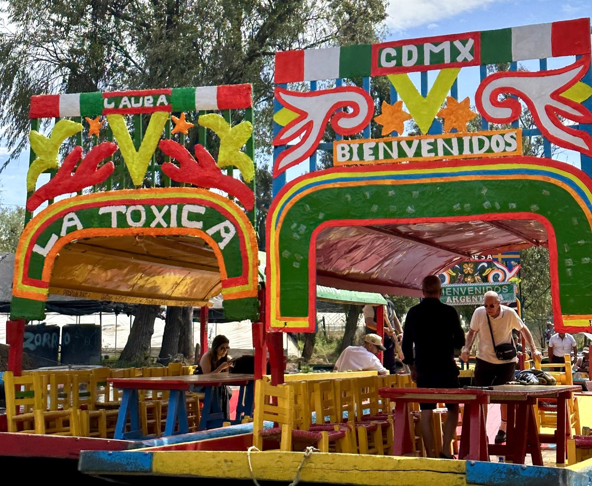 A view of people eating at colorful picnic tables with red, green, yellow and white signs overhead that read "La Toxica" and "CDMX Bienvenidos"
