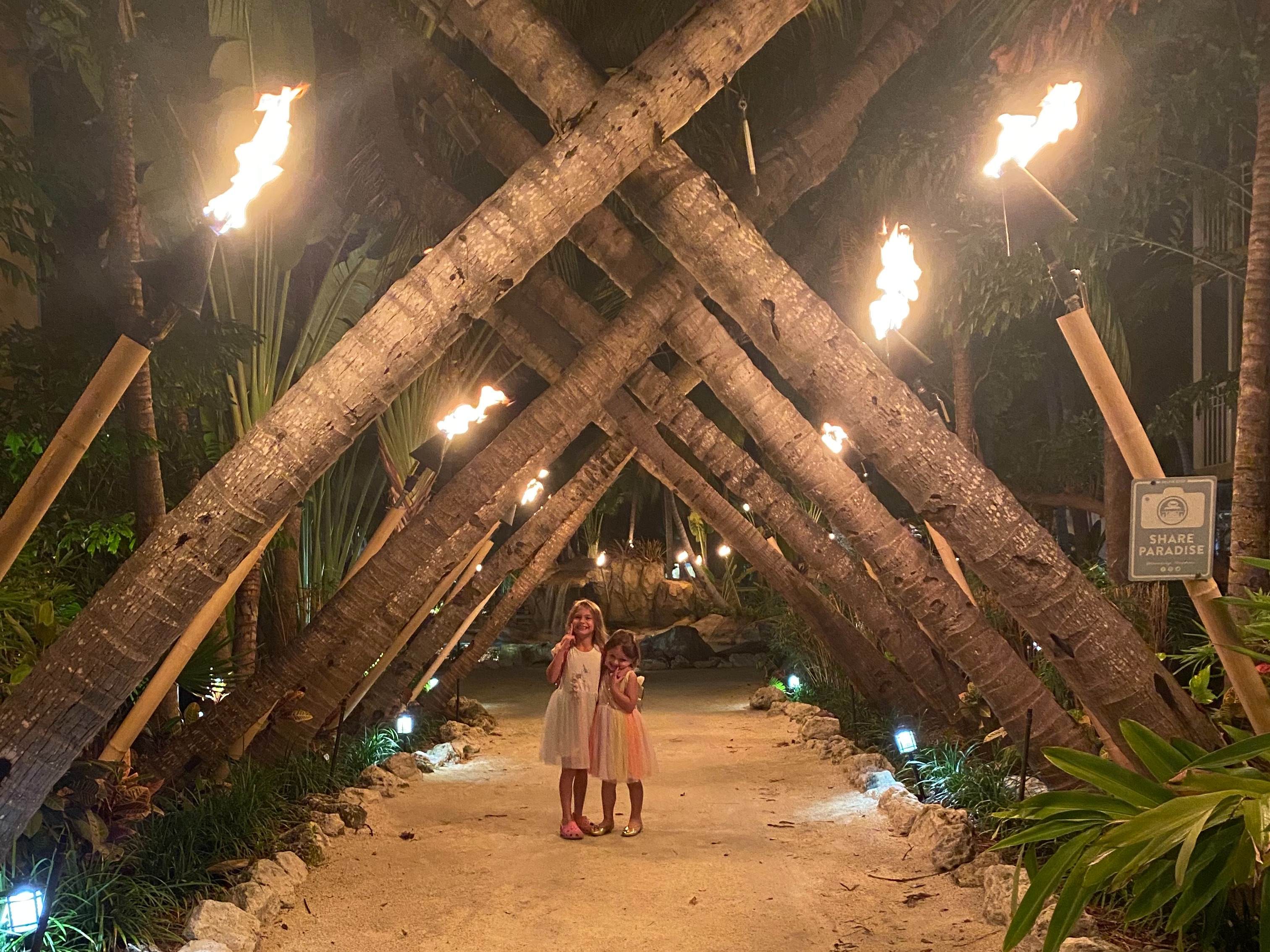 Kids are standing on a sandy pathway lined with lit torches and tropical vegetation at night.
