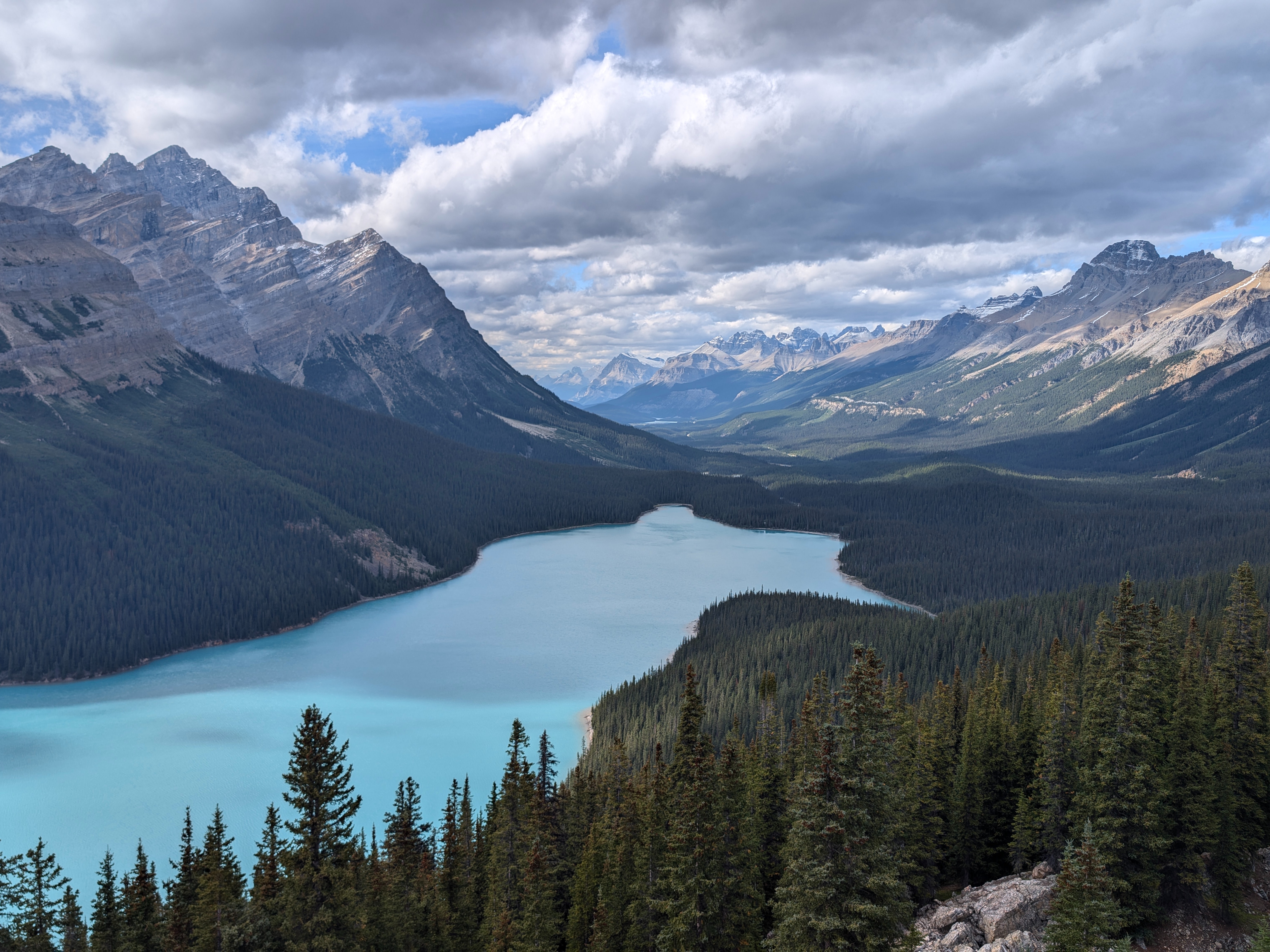 A panoramic view of a turquoise lake surrounded by dense forests and rugged mountains under a cloudy sky.