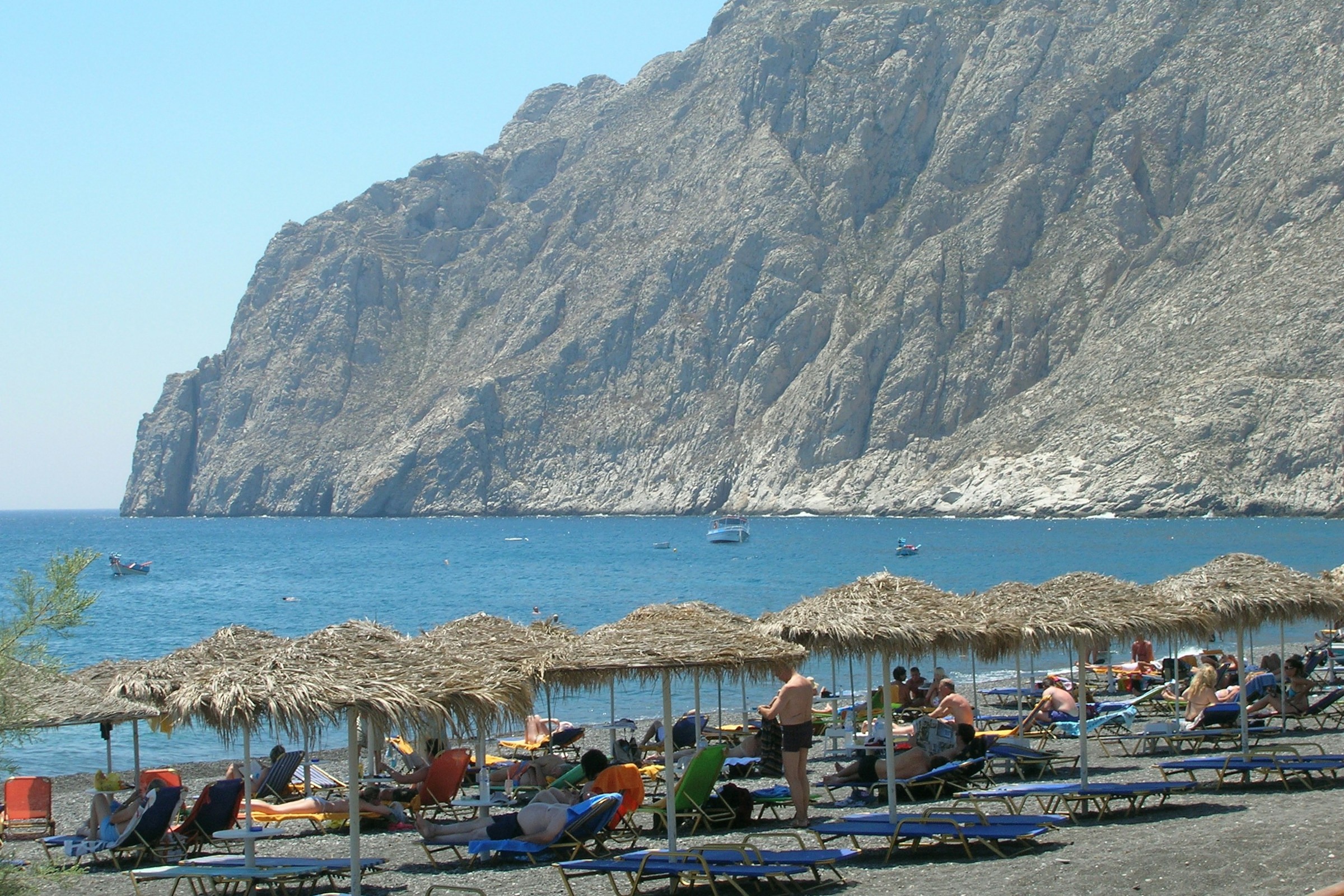 People in Kamari Black Sand Beach overlooking the calm water and a large mountain in the distance.