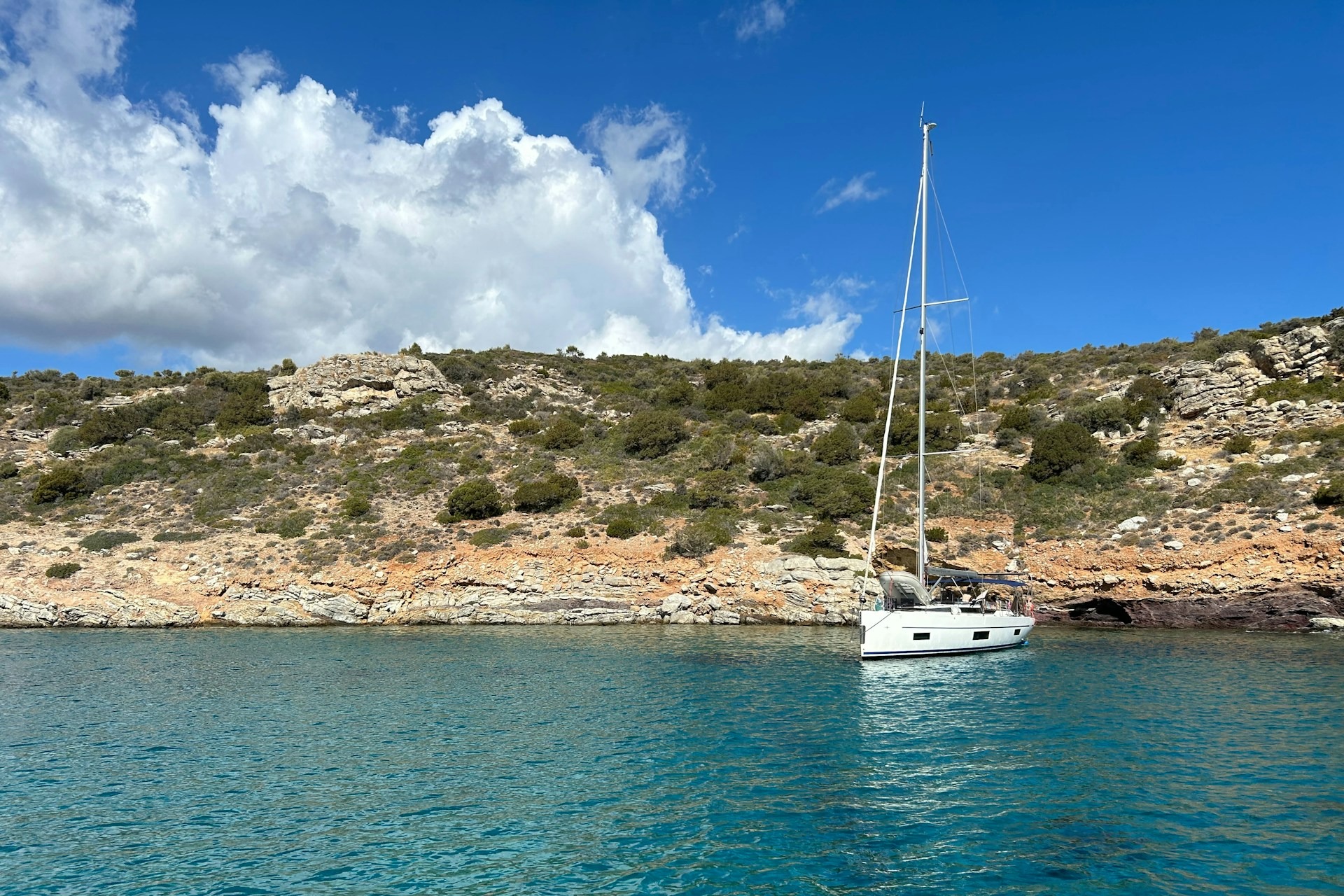 A view of a white sailboat floating along the shore of a rocky bank with green shrubs during the day.