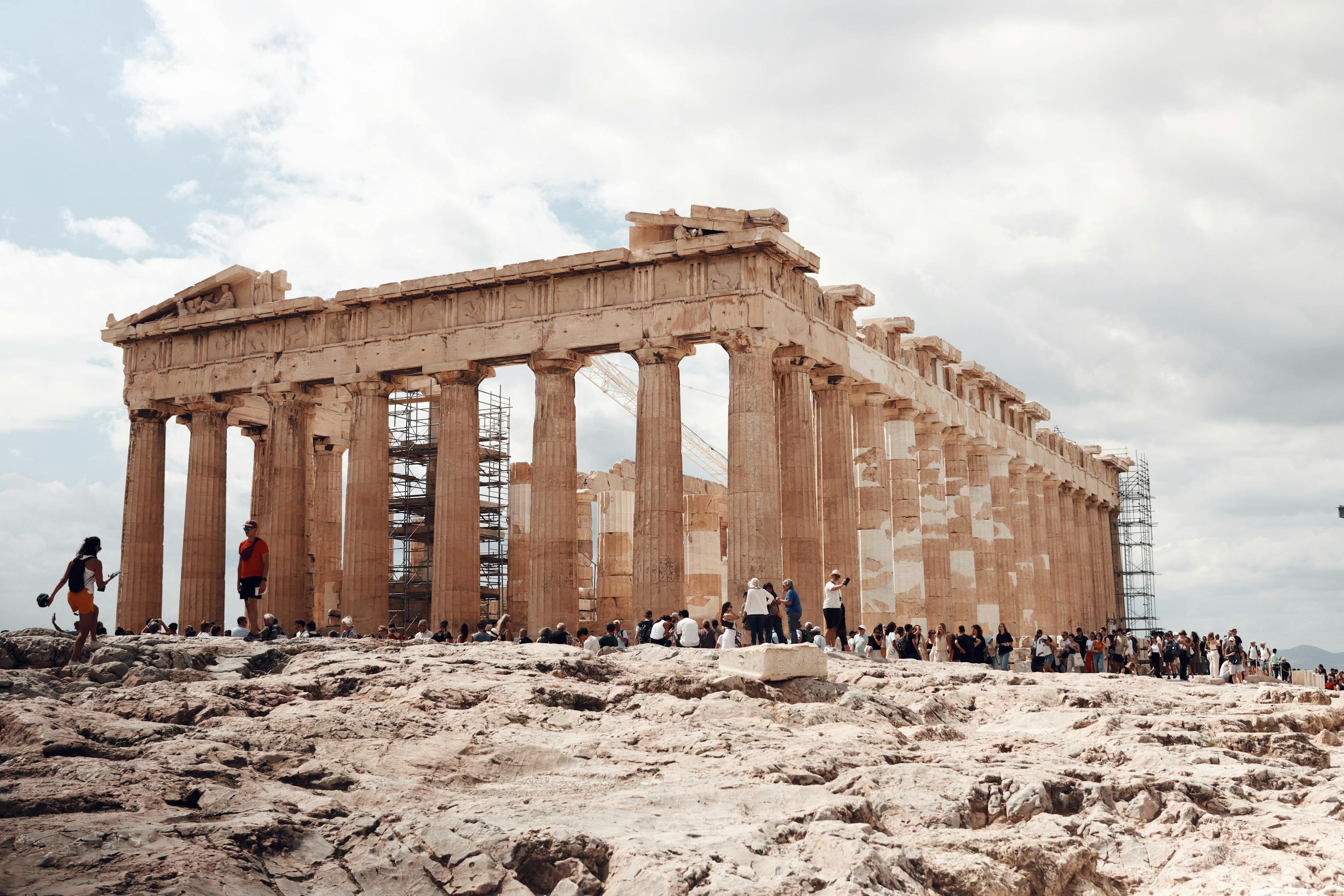 The Parthenon, an ancient Greek temple atop the Acropolis in Athens. Ancient white stone pillars and tourists surrounding it.