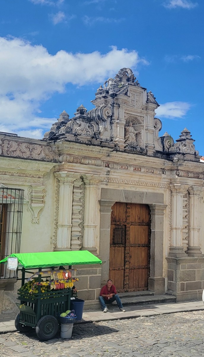 A man sits in the shade of an historic building next to his cart selling fruit and wares on a sunny day with scattered clouds.