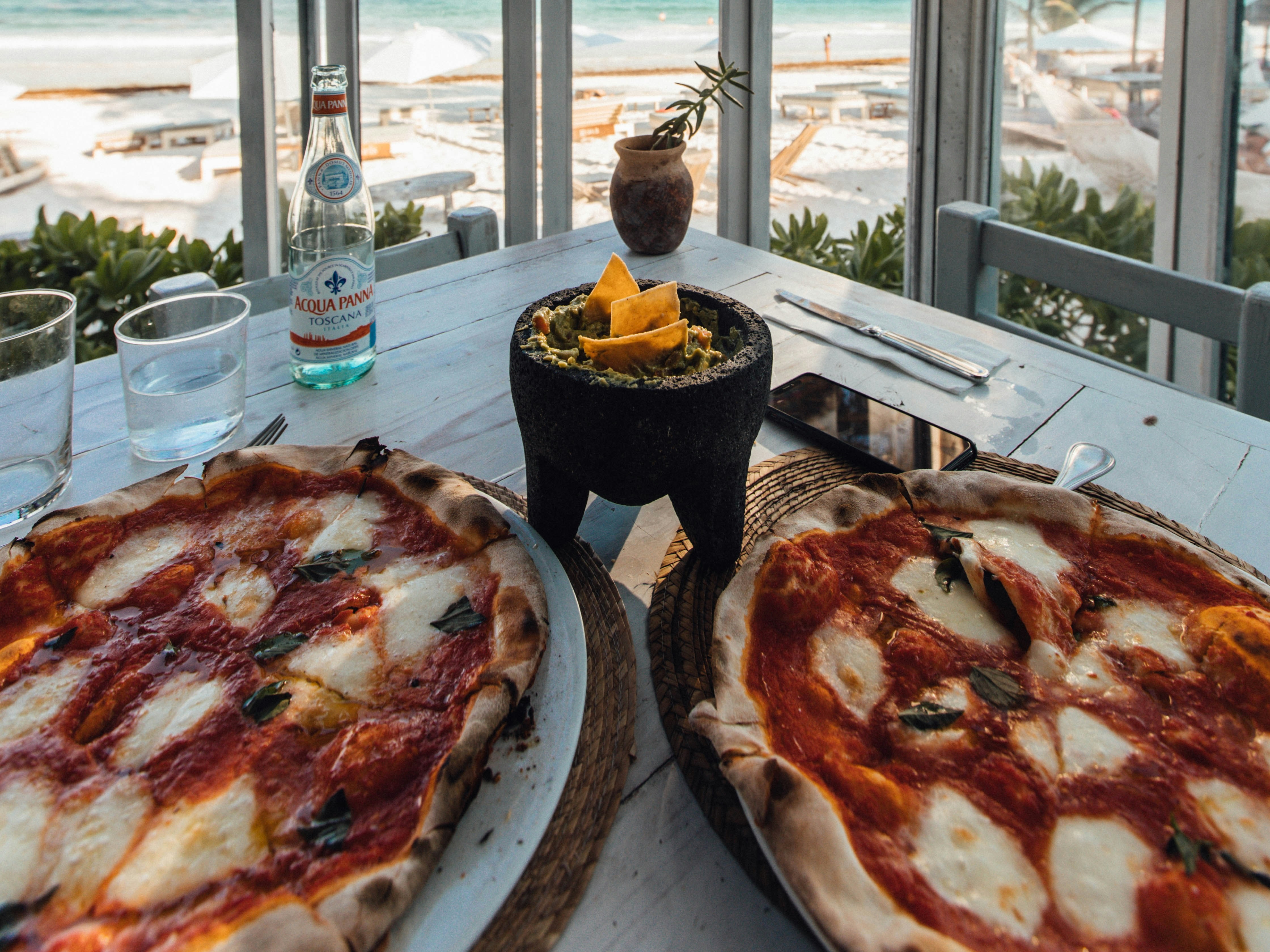 A scenic beach view from a window, with two pizzas and a drink on the sill creates an inviting dining atmosphere.