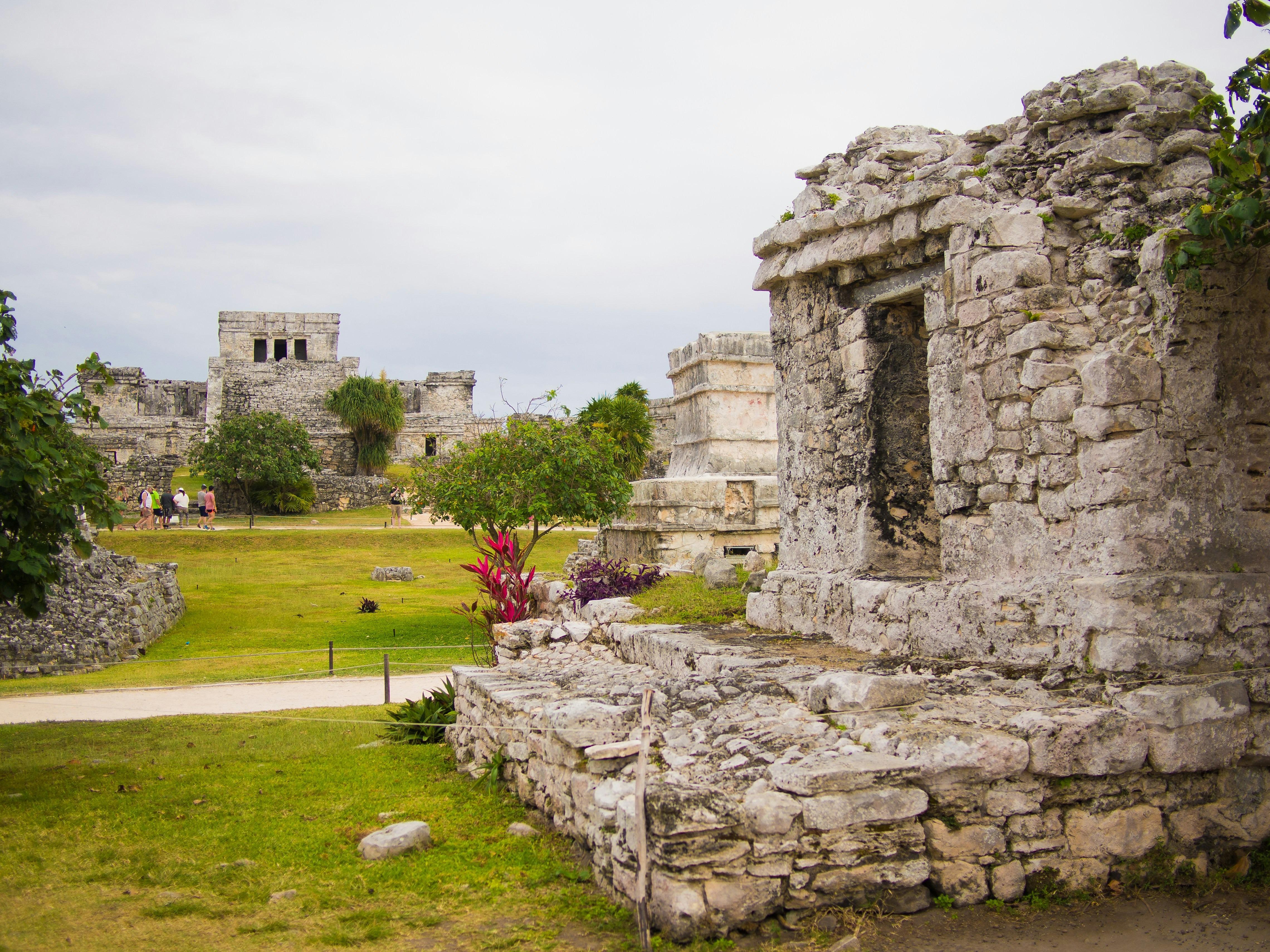 Ancient stone ruins stand amidst a grassy field, with visitors exploring under a cloudy sky.