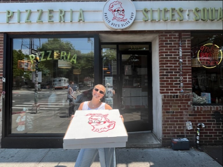 A person holding a pizza box in front of a pizzeria.