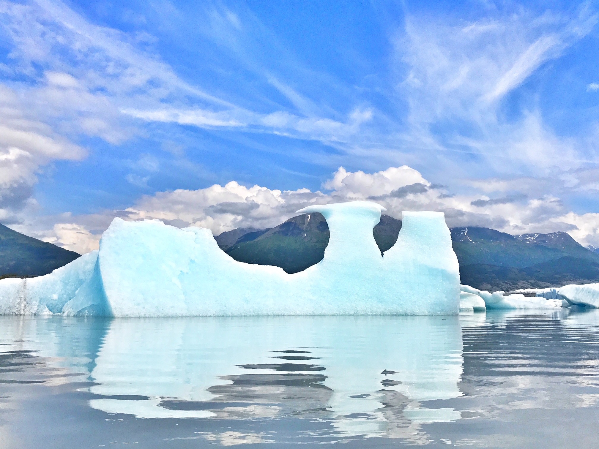 A large, sculptural iceberg with arches is reflected in calm water, set against a mountainous backdrop.