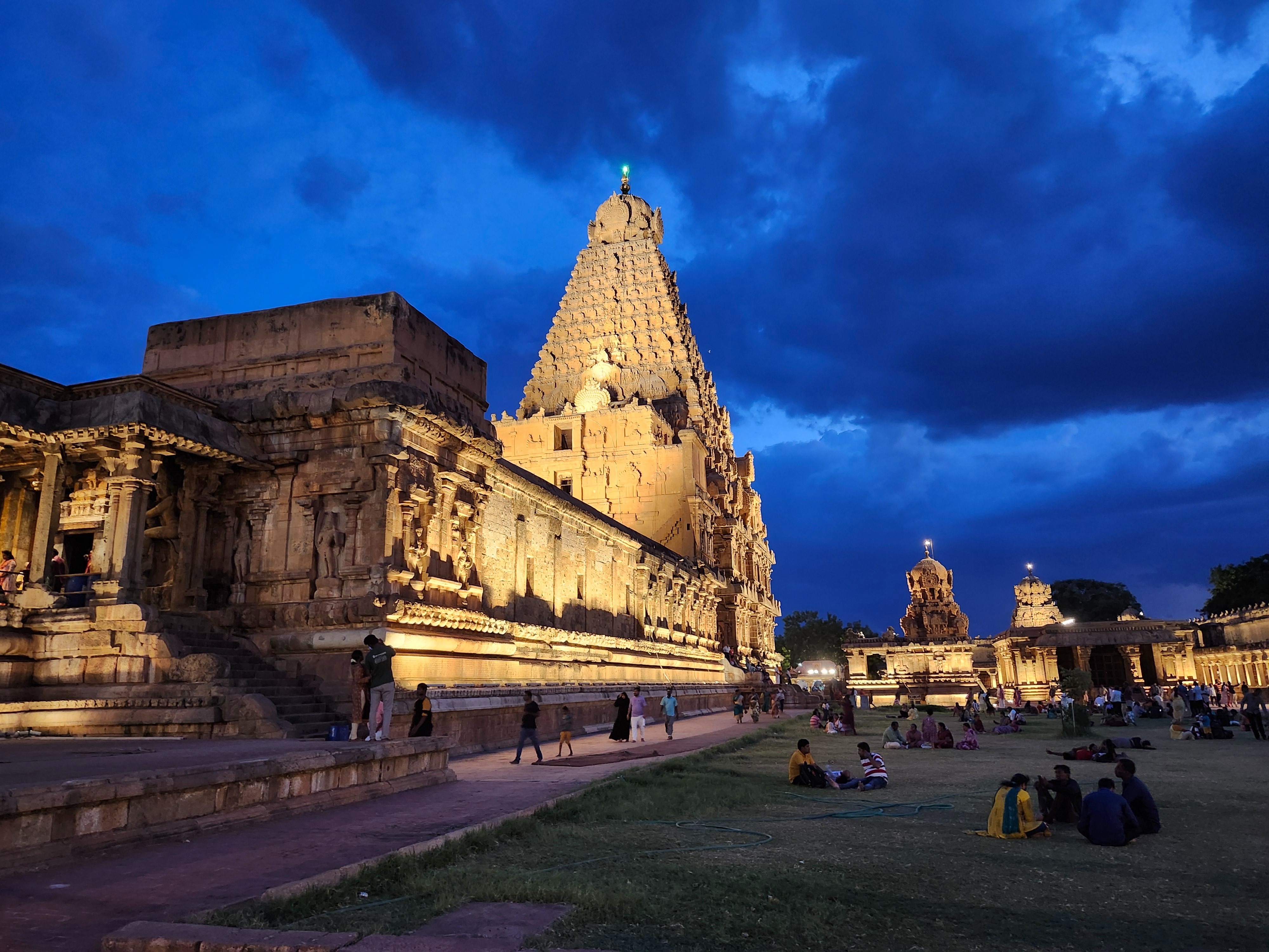 The illuminated Brihadeeswarar Temple at dusk, with people relaxing on the grass in the foreground.