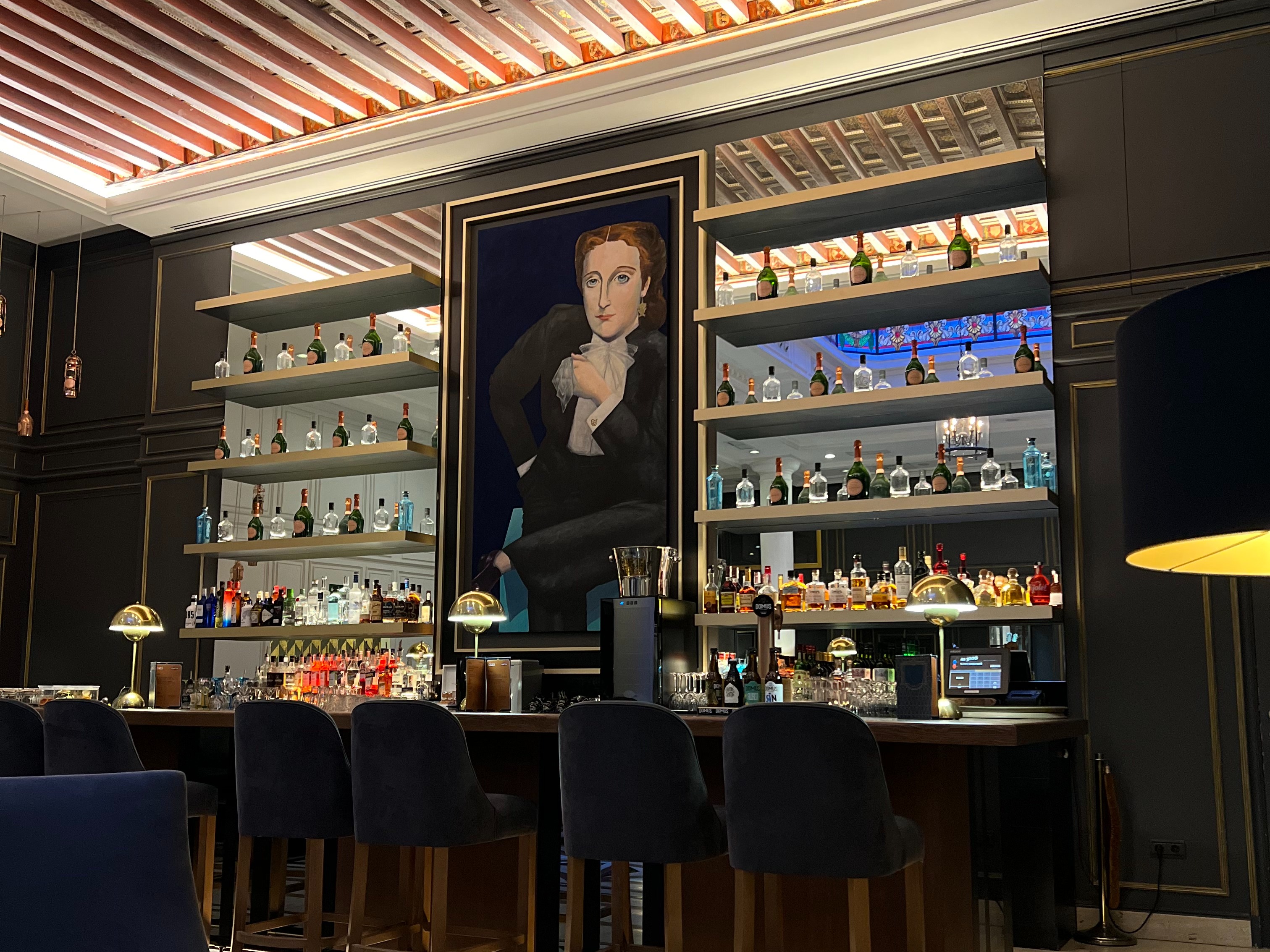 An interior space, possibly a bar or restaurant, with shelves of bottles, artwork on the wall, and seating areas.
