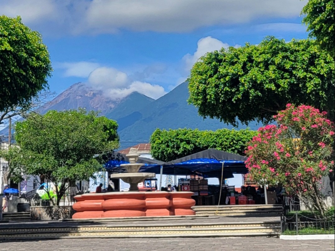 A vibrant outdoor scene with green trees, red flowers, blue umbrellas, benches, and mountains in the background under a blue sky with fluffy clouds.
