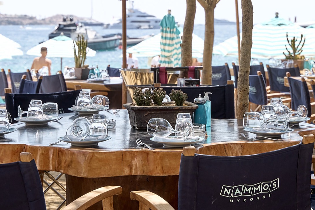 A large, round dining table overlooks the sea as boats cruise by on a sunny day at Nammos Mykonos.