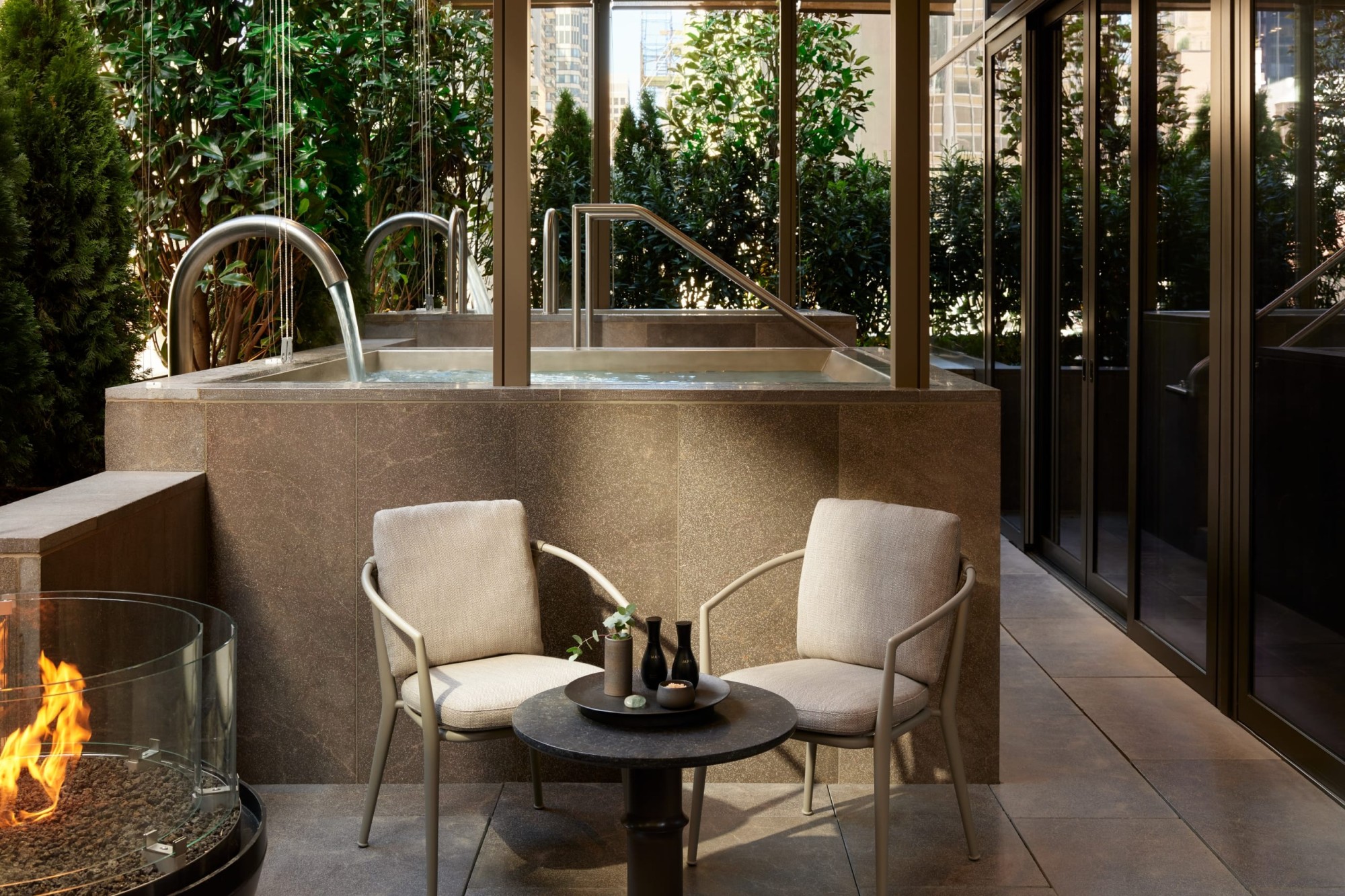 Private outdoor space with a hot tub, a fire pit and a small table setting; all surrounded by potted plants for privacy
