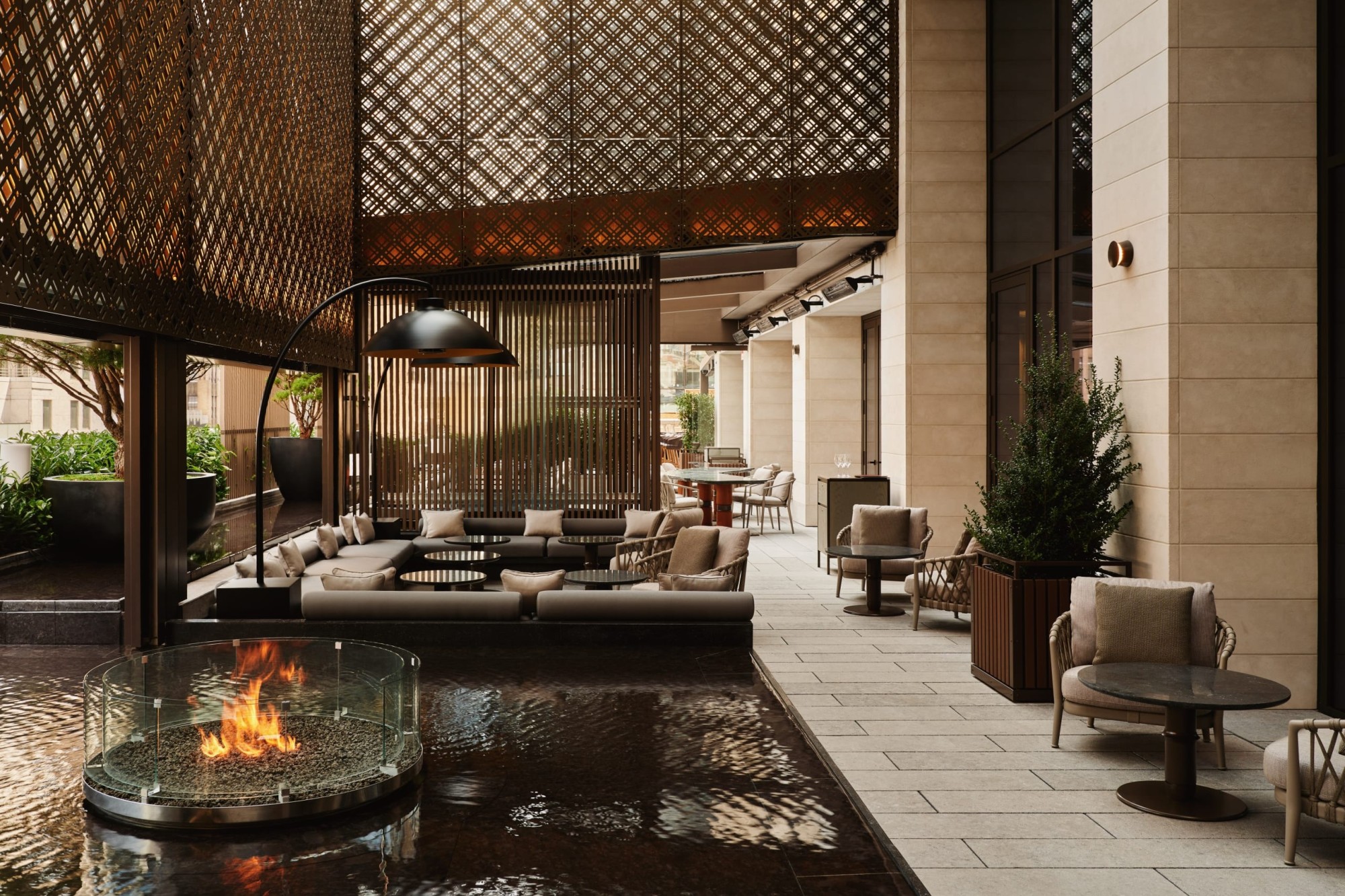 A water feature with a firepit at the center is featured prominently on a terrace, which also hosts lounge seating and potted plants