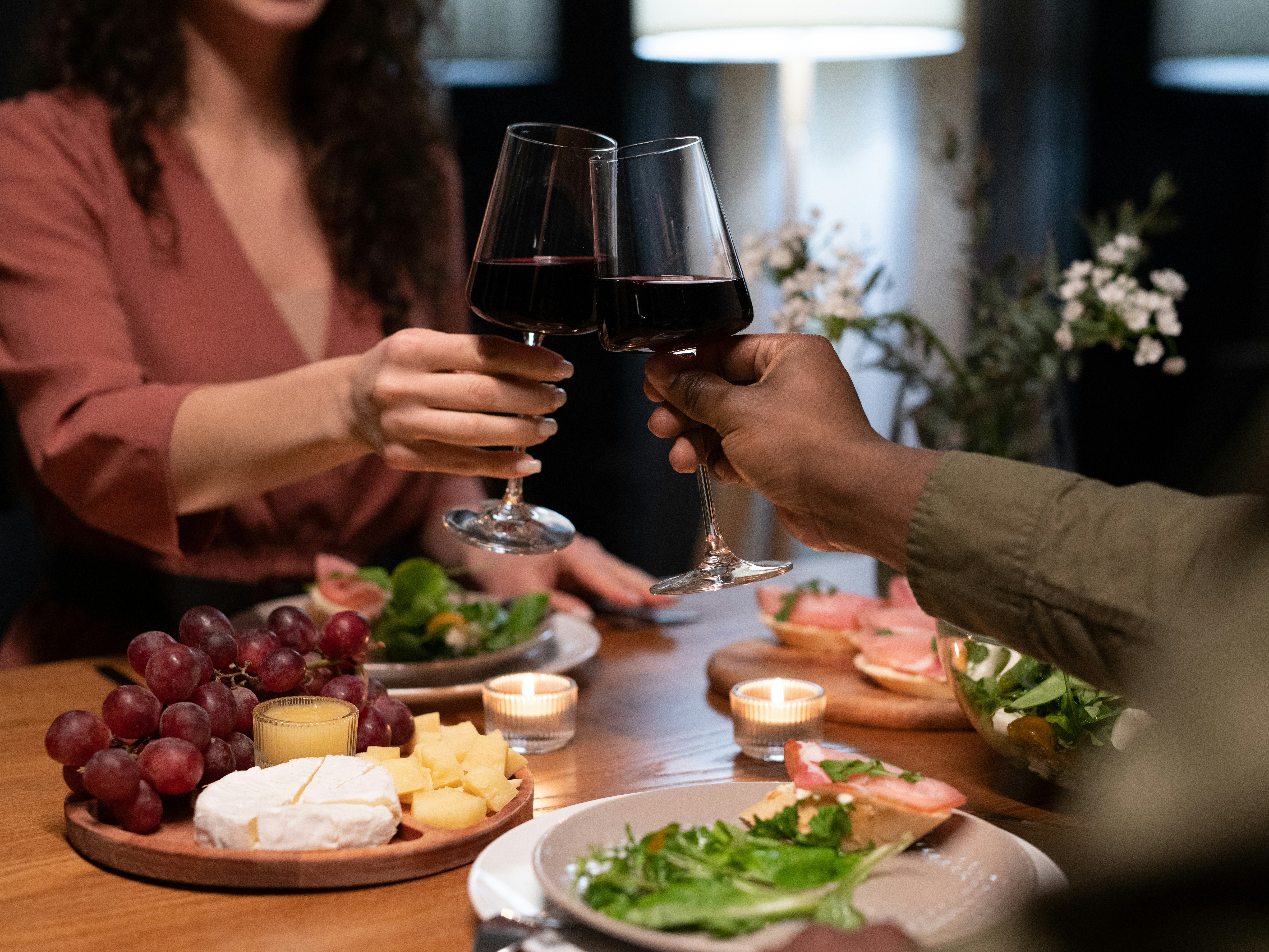 Two individuals toast with glasses of red wine over a candlelit dinner table.