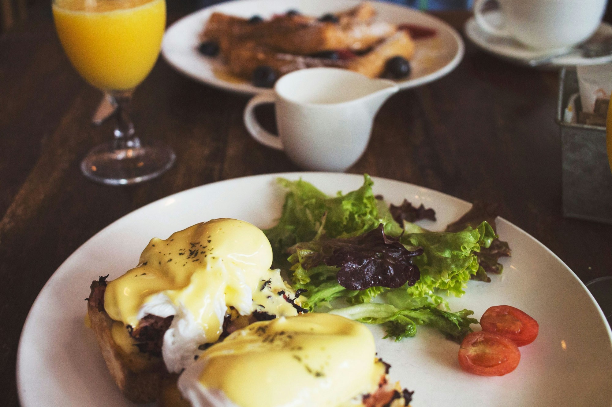A food view of eggs benedict on a white plate with greens and tomato garnish and a glass of orange juice.