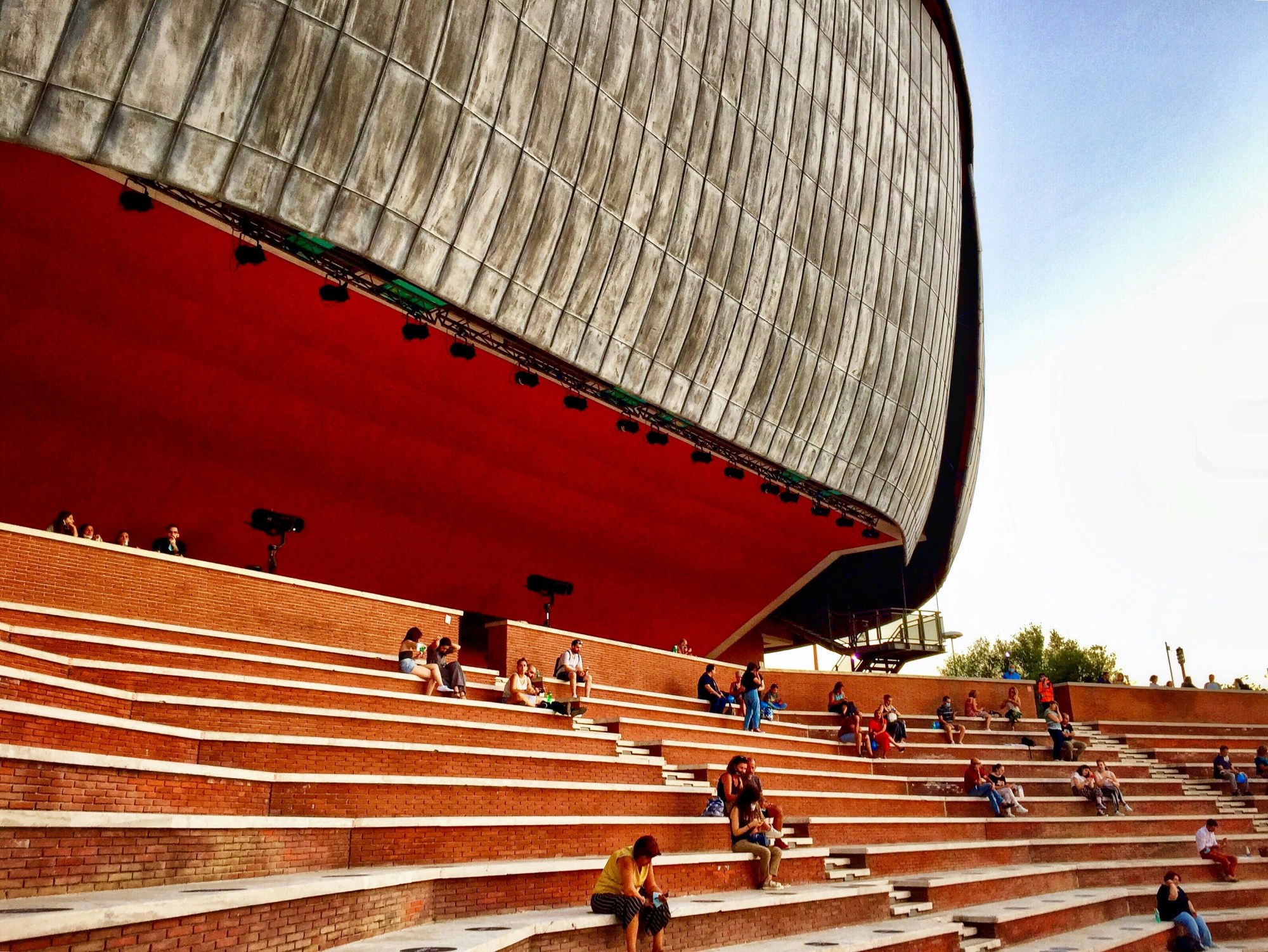 People sitting in a large stadium with orange bleacher seating during the day.