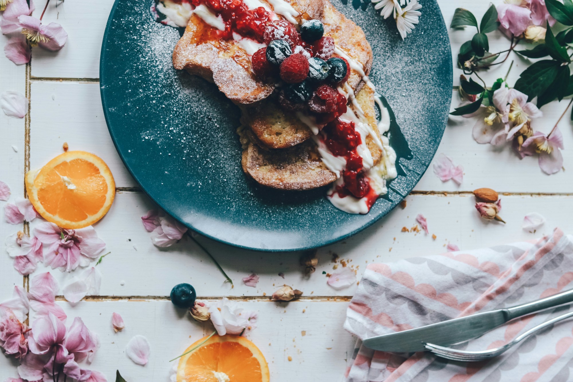 A food view of gourmet french toast topped with mixed berries and cream, on a blue plate surrounded by flowers and citrus garnish.