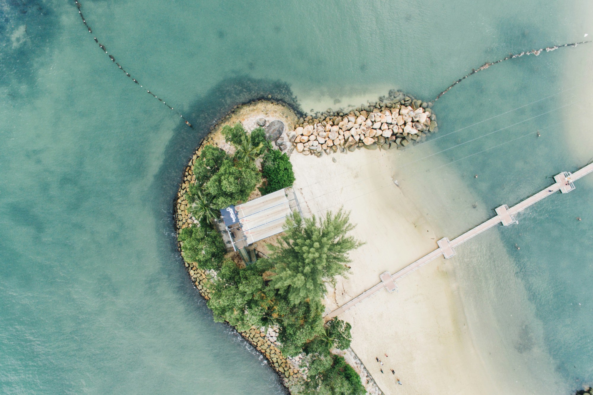 An aerial view of Sentosa Island, with white sand beaches, white rocks, palm trees and a clear blue ocean surrounding it.