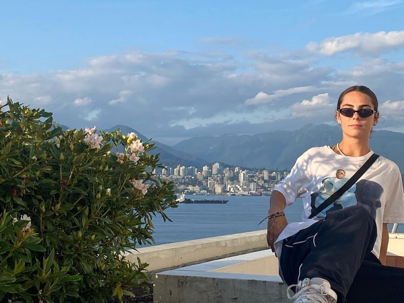 An individual sits on a ledge, overlooking a cityscape and mountains under a clear sky with a bird flying by.