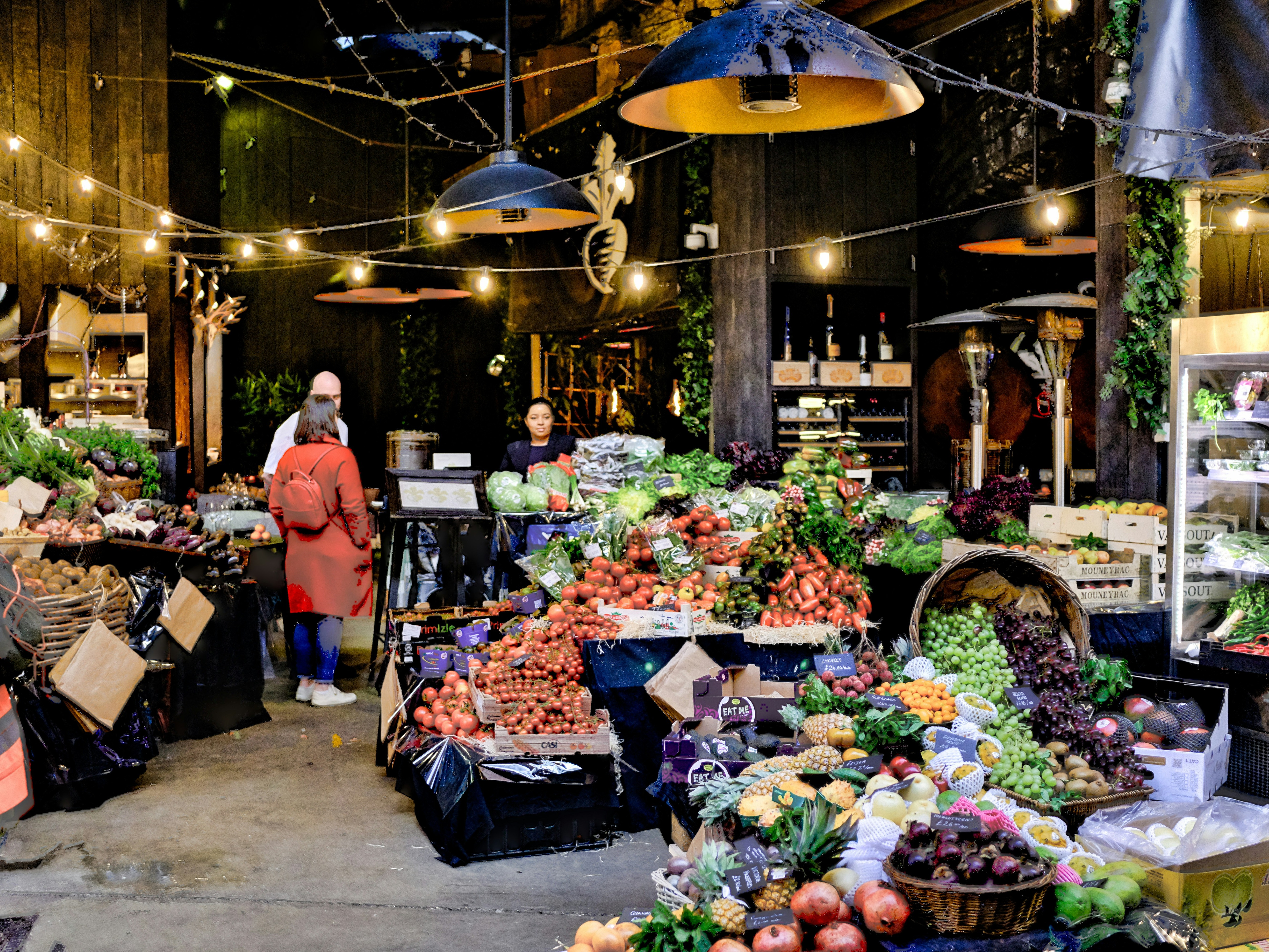 The image depicts a vibrant indoor market with an array of fresh produce and a person in an orange coat.