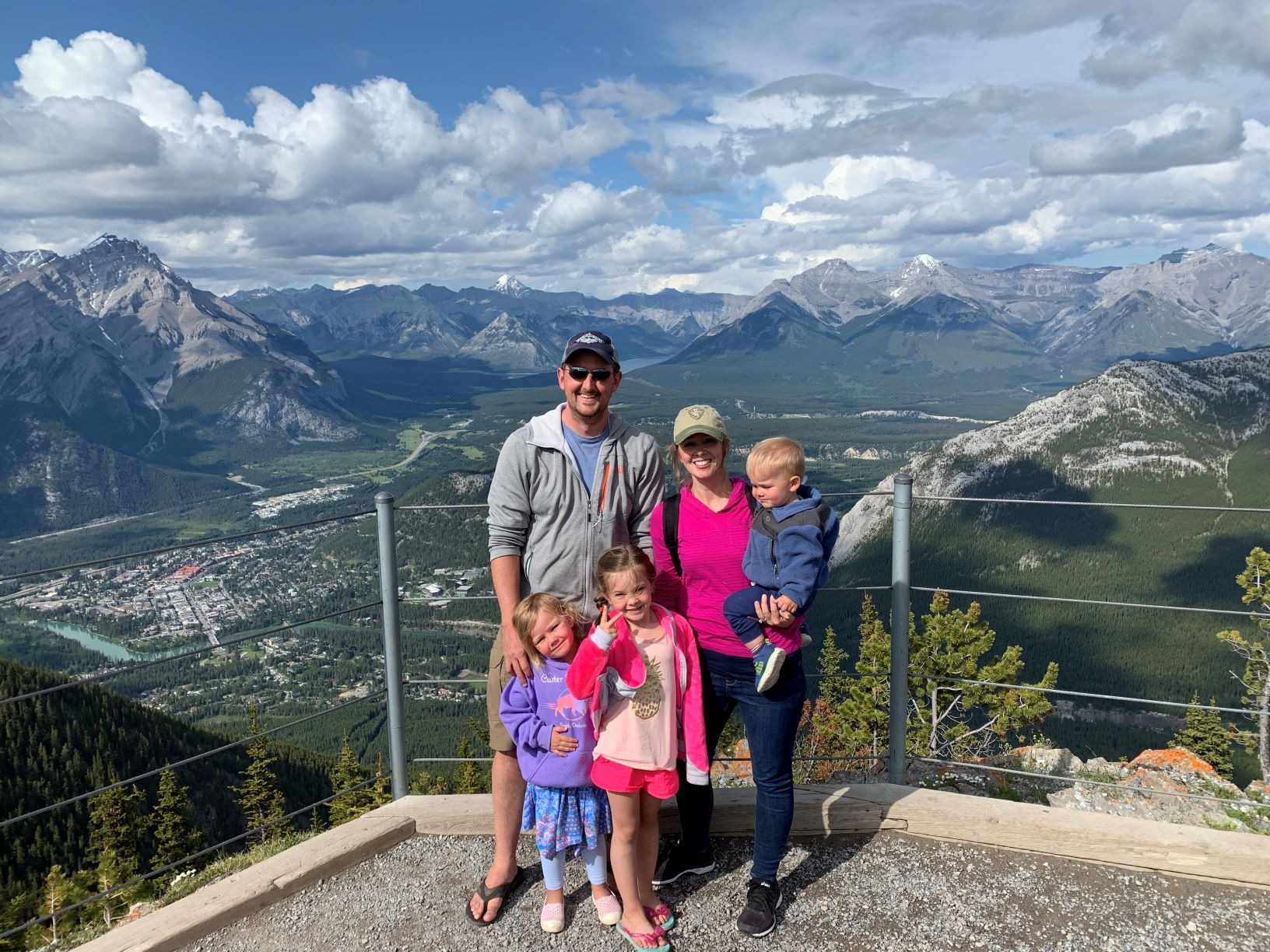A group of people stand on an observation platform with a panoramic view of a mountainous landscape and a town below.