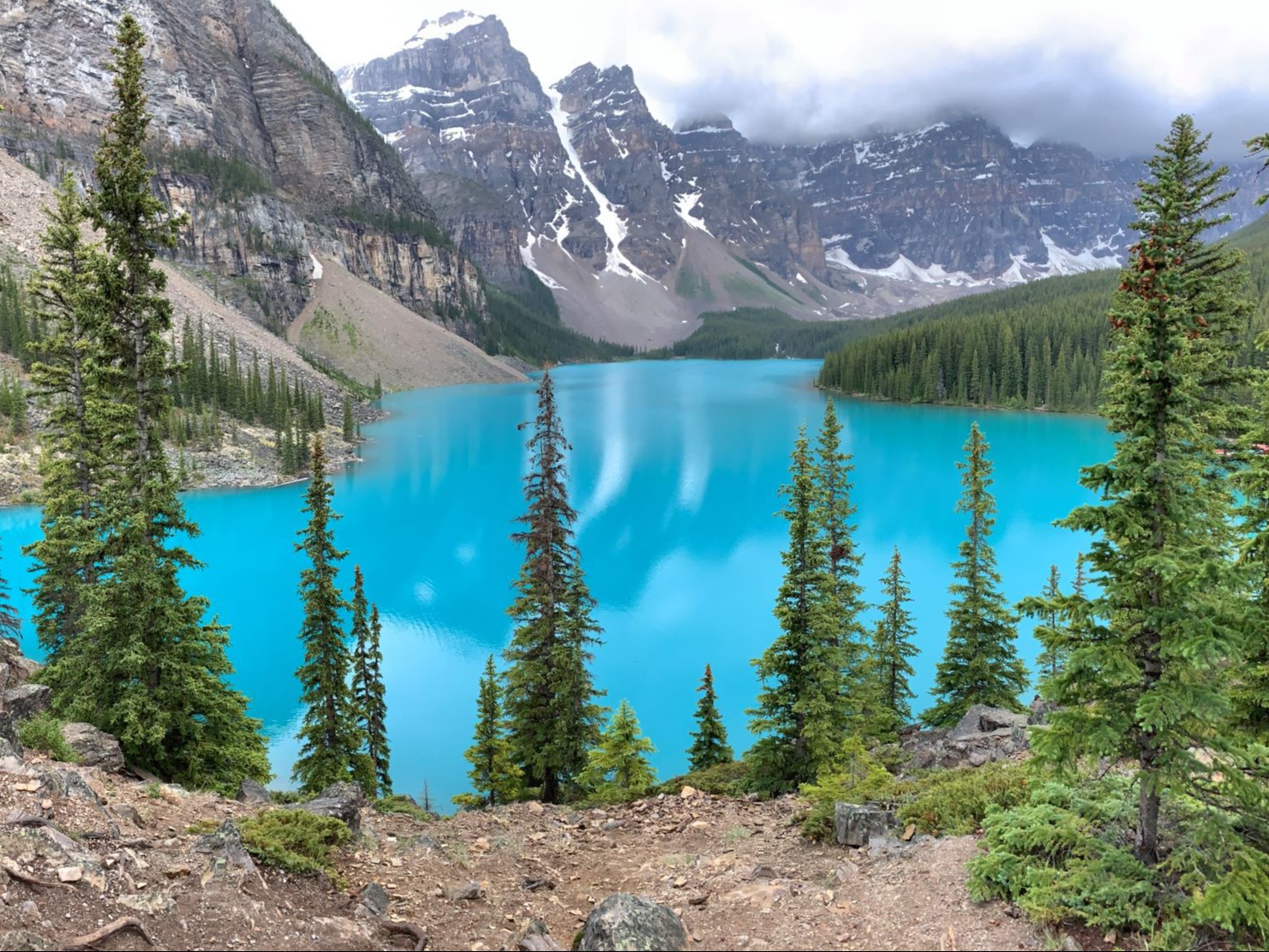 Forested areas and rocky mountains surround a vibrant turquoise lake under a cloudy sky.