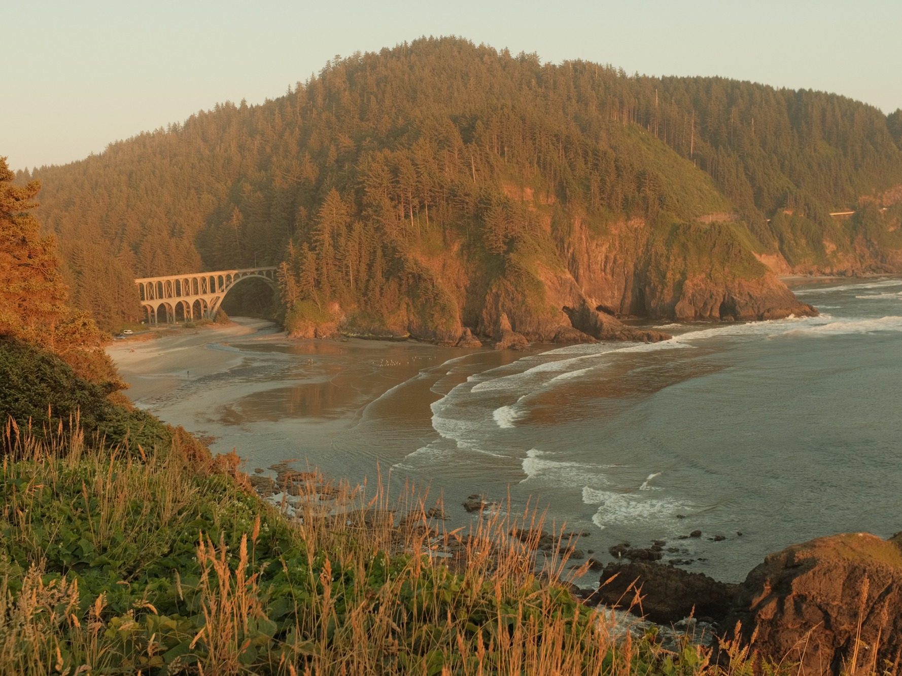 A picturesque coastal scene featuring a bridge, forested cliffs, and a sunlit beach.
