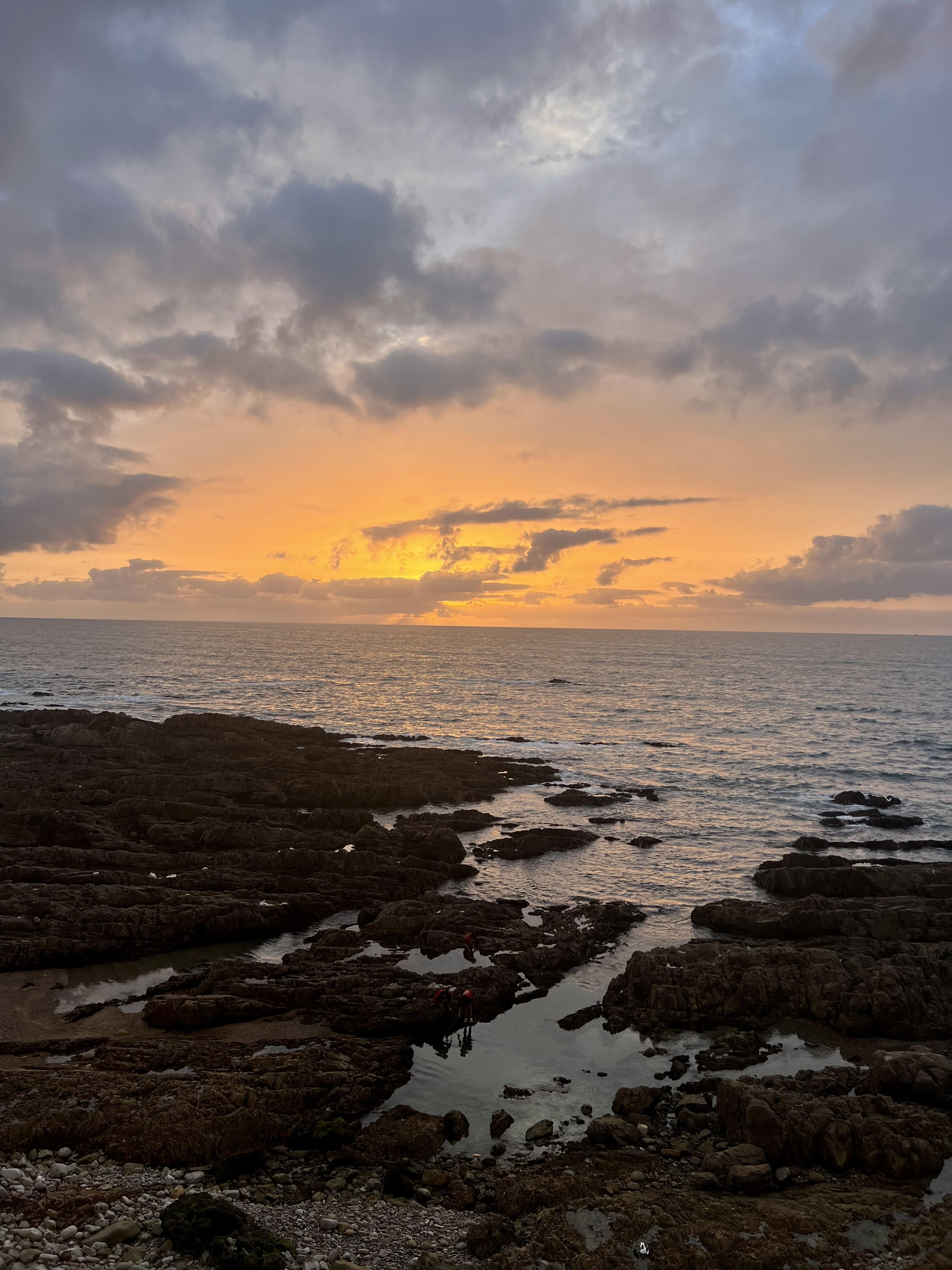 The sunset sets the sky on fire as the sun disappears behind the clouds and waves lap the rocky shore.