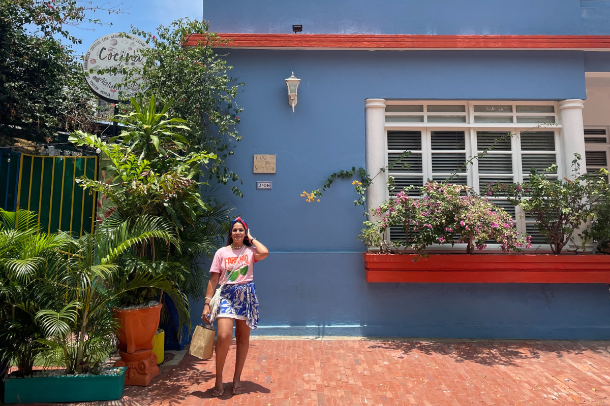 The advisor standing in front of a blue building with red trim and flower pots on the window sill and plants next to the building. There is a sign that reads "Cocina de Emy Natural Food" restaurant in Santa Marta.