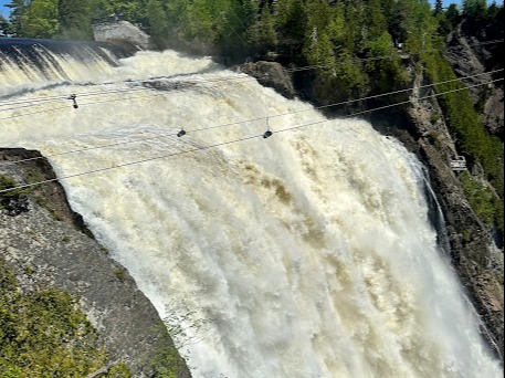 A large waterfall with a suspension bridge above and multiple zip lines crossing in front of it.