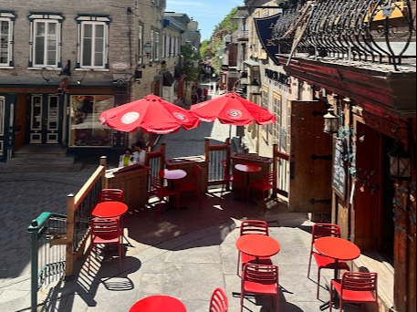 A sunny street view with outdoor red tables and umbrellas, flanked by old buildings, suggests a quaint European-style cafe setting.