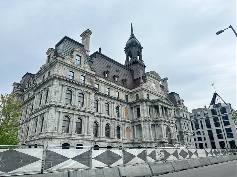 The image shows a large, ornate building with classical architecture, featuring multiple levels, a variety of windows, and a central tower under an overcast sky.
