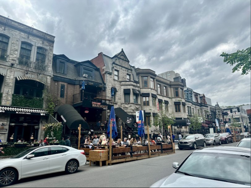 A lively street scene with outdoor dining, cars, and historic buildings under an overcast sky.