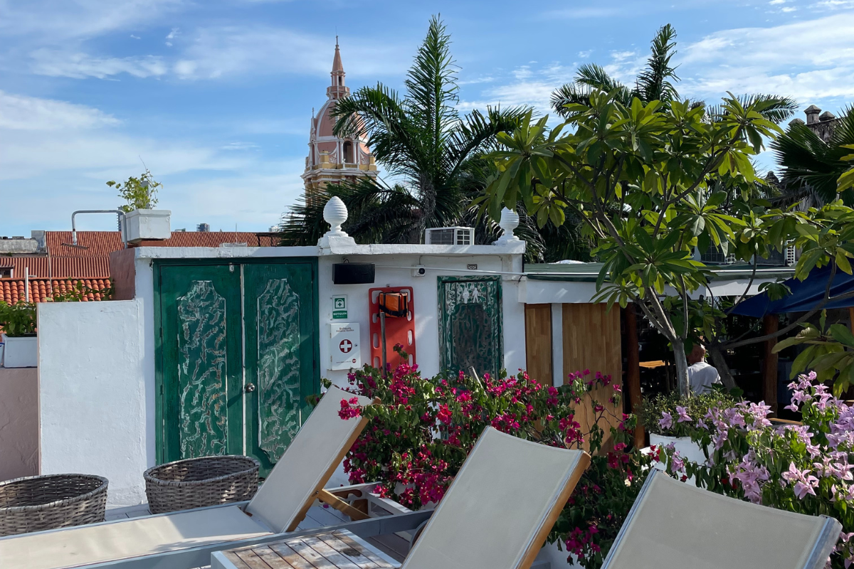 A view of lounge chairs on a patio with flowers, palm trees and a small white building that has decorated green doors. In the distance there is a tall tower with a dome.