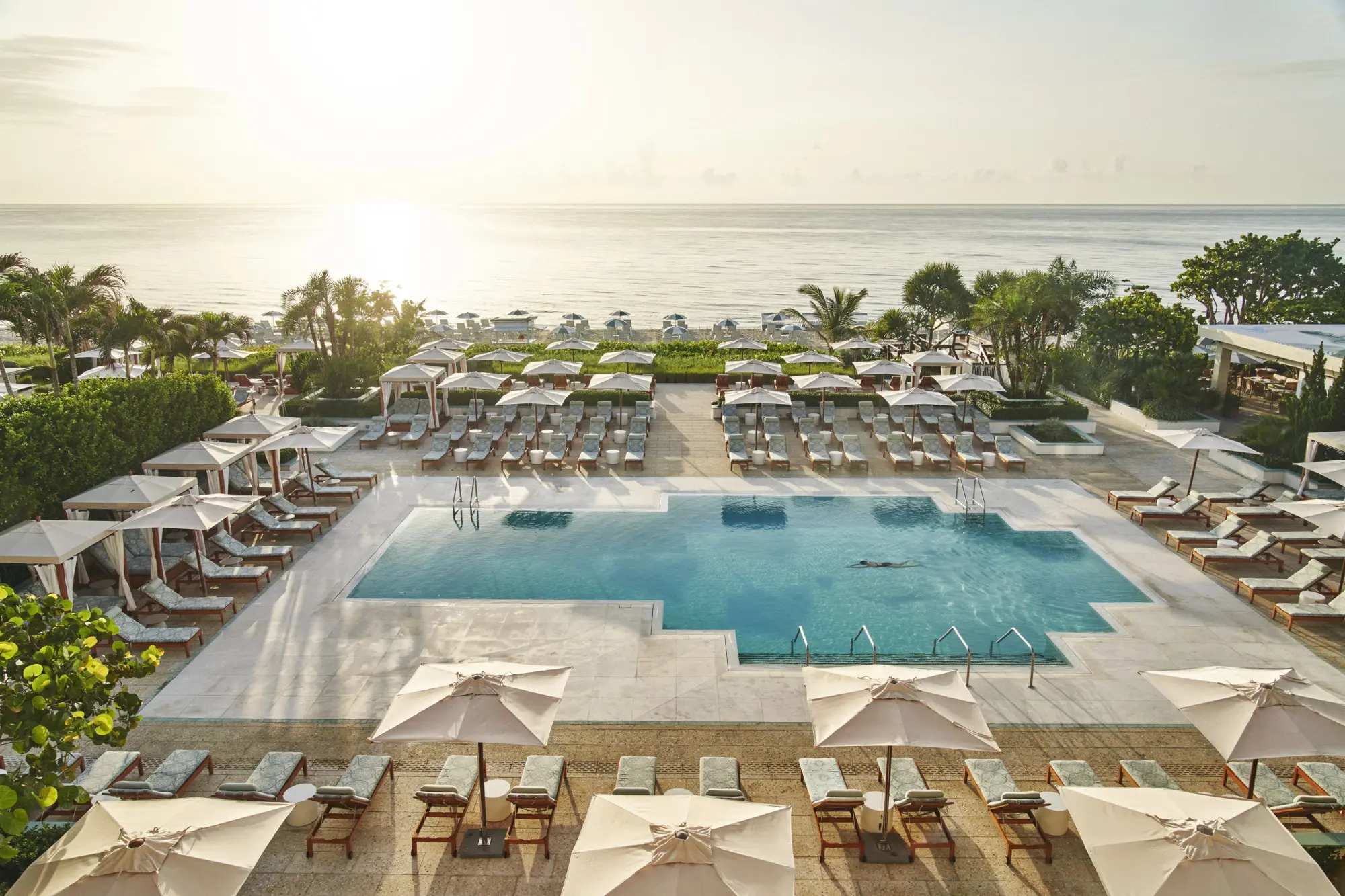 The sun casts a small glare on the camera as it rises: loungers and potted plants surround a luxurious pool area, with the ocean visible in the distance