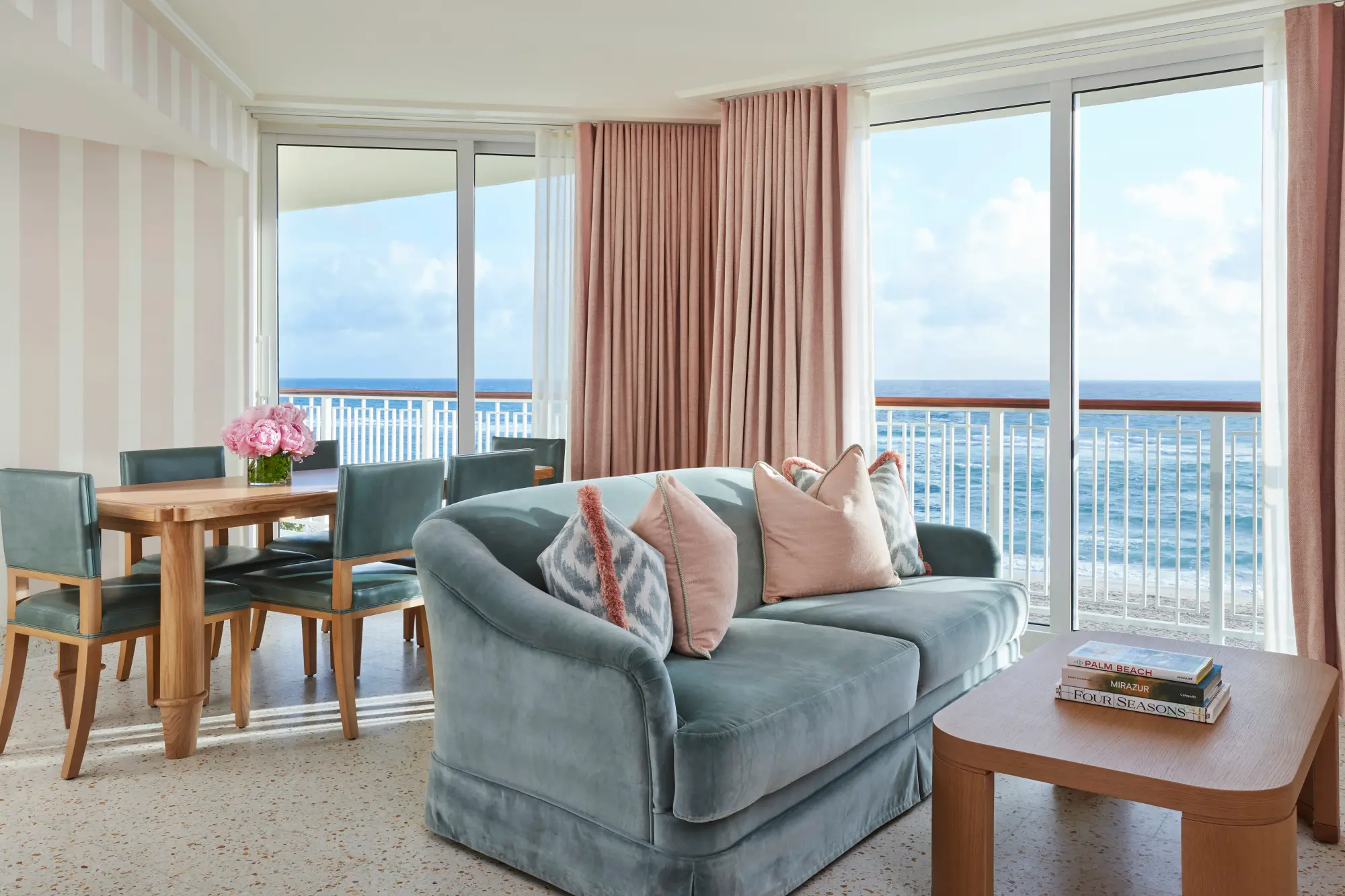 Coastal-themed furniture fills a living space with huge windows and a sliding glass door that leads to a private balcony overlooking the ocean