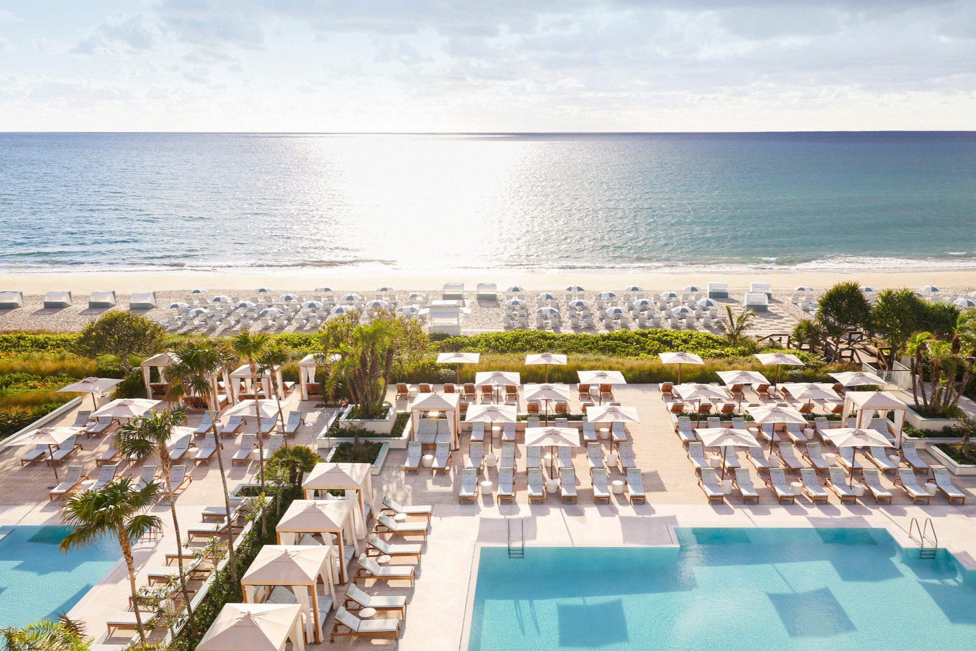 Rows of loungers and cabanas line large pools before manicured gardens that lead to a private beach with calm waves