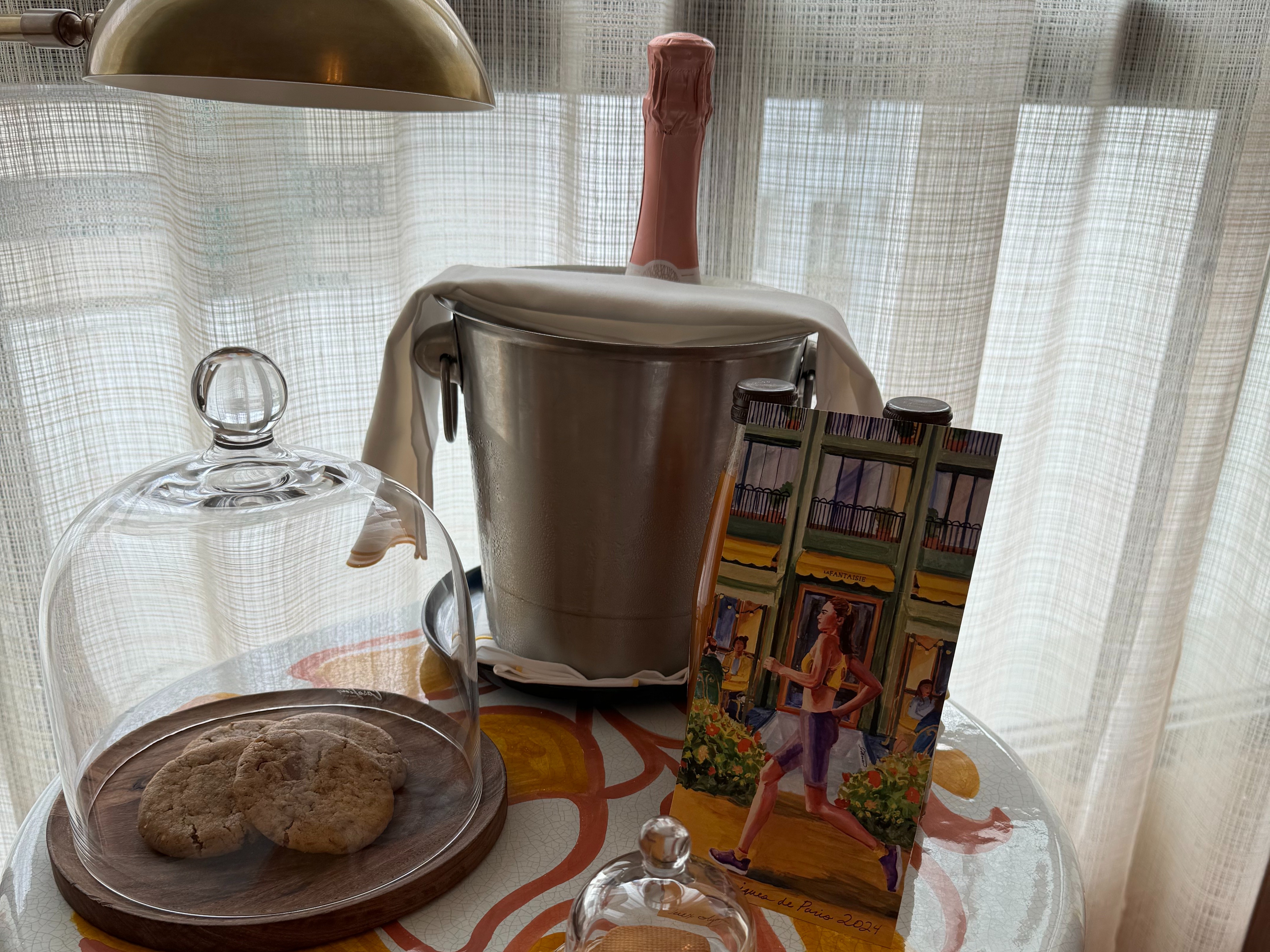 An image of a welcome amenity at La Fantaisie hotel in Paris with a bottle of champagne in a bucket, a vintage card and some sweet treats, displayed on a small round table.