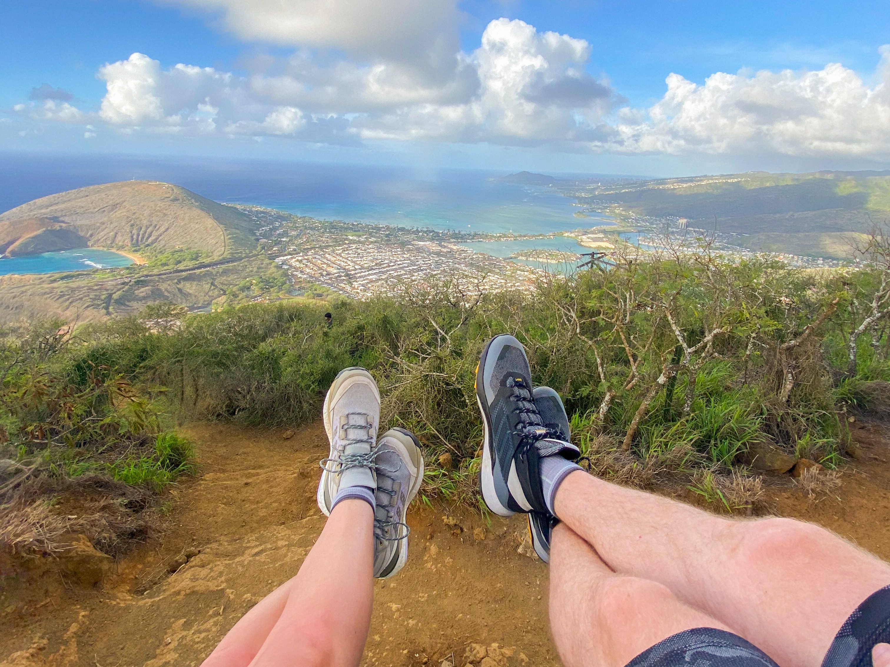 A view of two people's feet at a very high peak of a mountain with an amazing view below.
