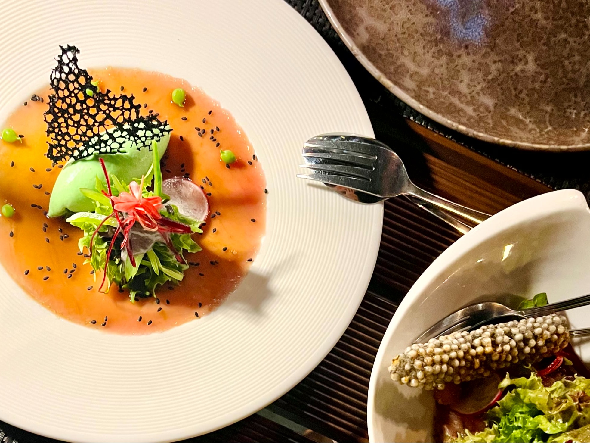 The image shows a close-up of a gourmet dish with vibrant colors and intricate plating, accompanied by cutlery and another dish partially visible.