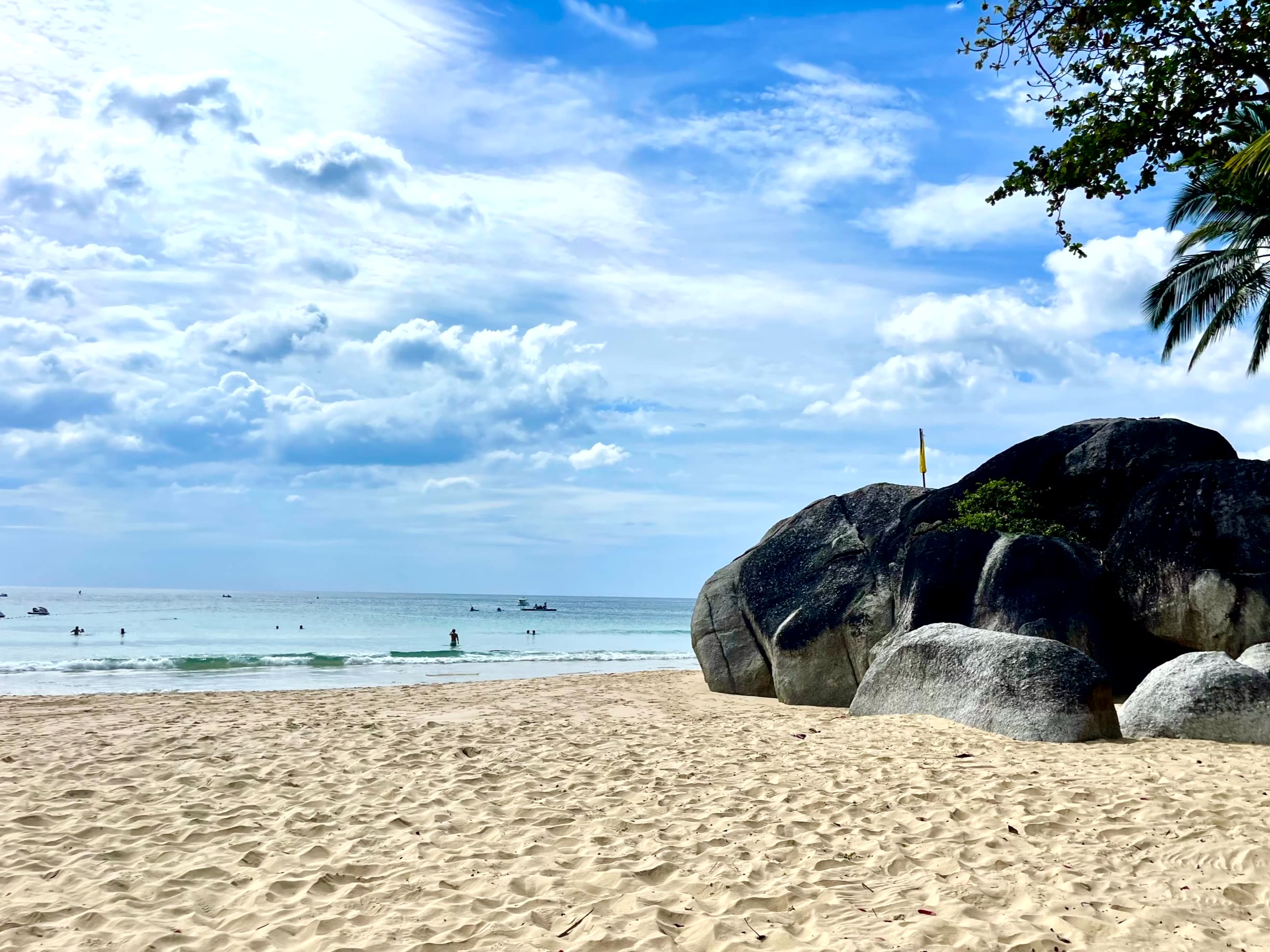The image shows a serene beach with large rocks, palm trees and people enjoying the sea under a partly cloudy sky.