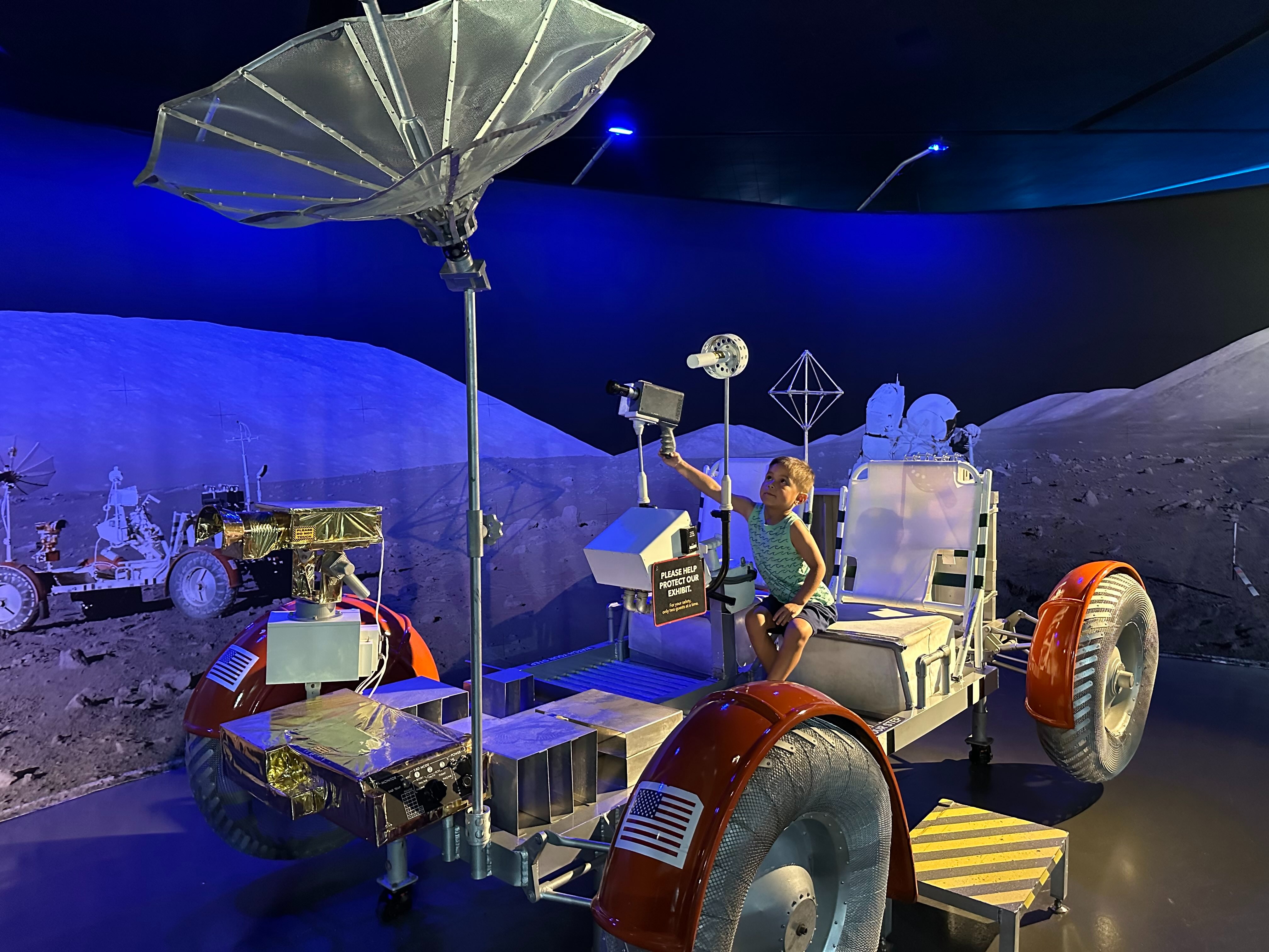 The image shows a display of a lunar rover model with various scientific instruments, set against a backdrop simulating the moon’s surface.